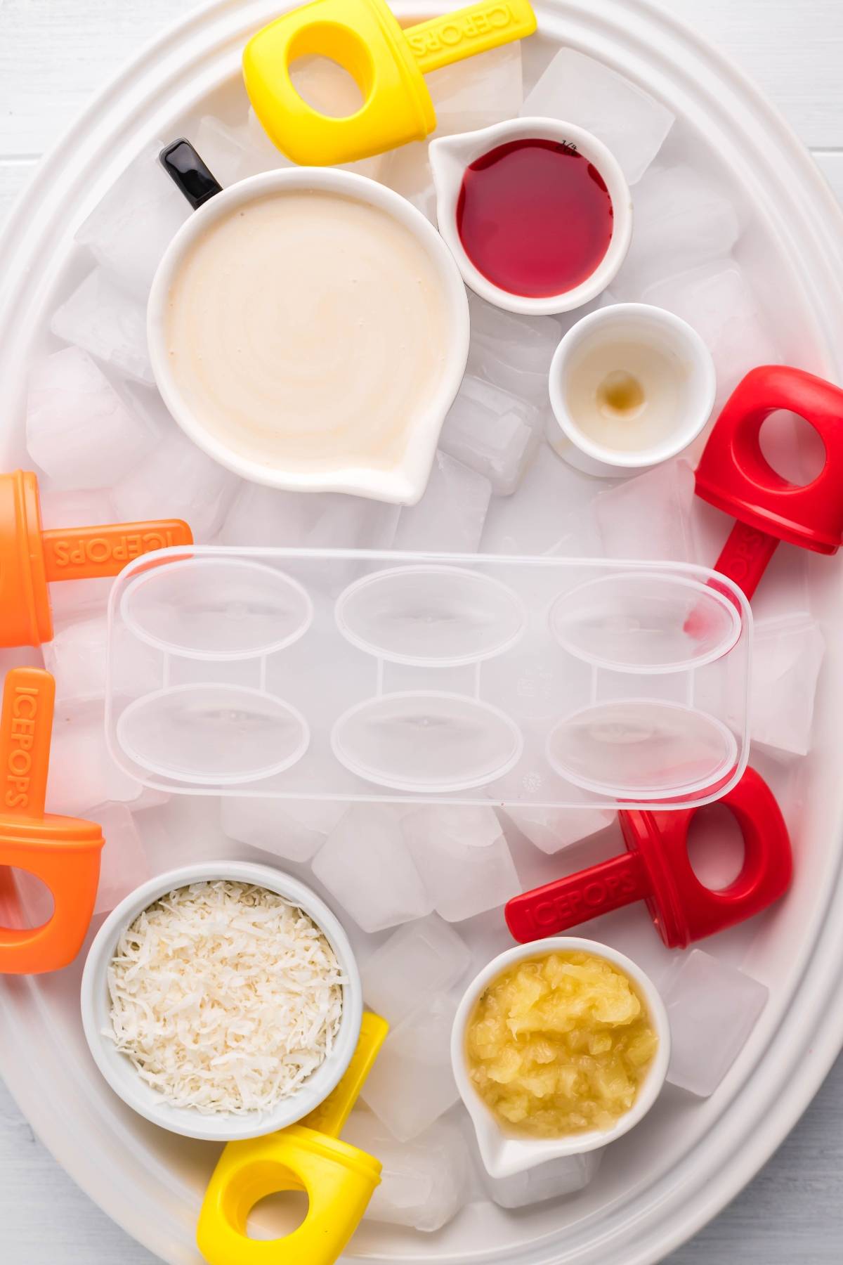 An oval tray with ice, bowls of coconut milk, pineapple, shredded coconut, juice, and popsicle molds—perfect for making Sugar Free Popsicles or refreshing Tropical Sunrise Popsicles at home.