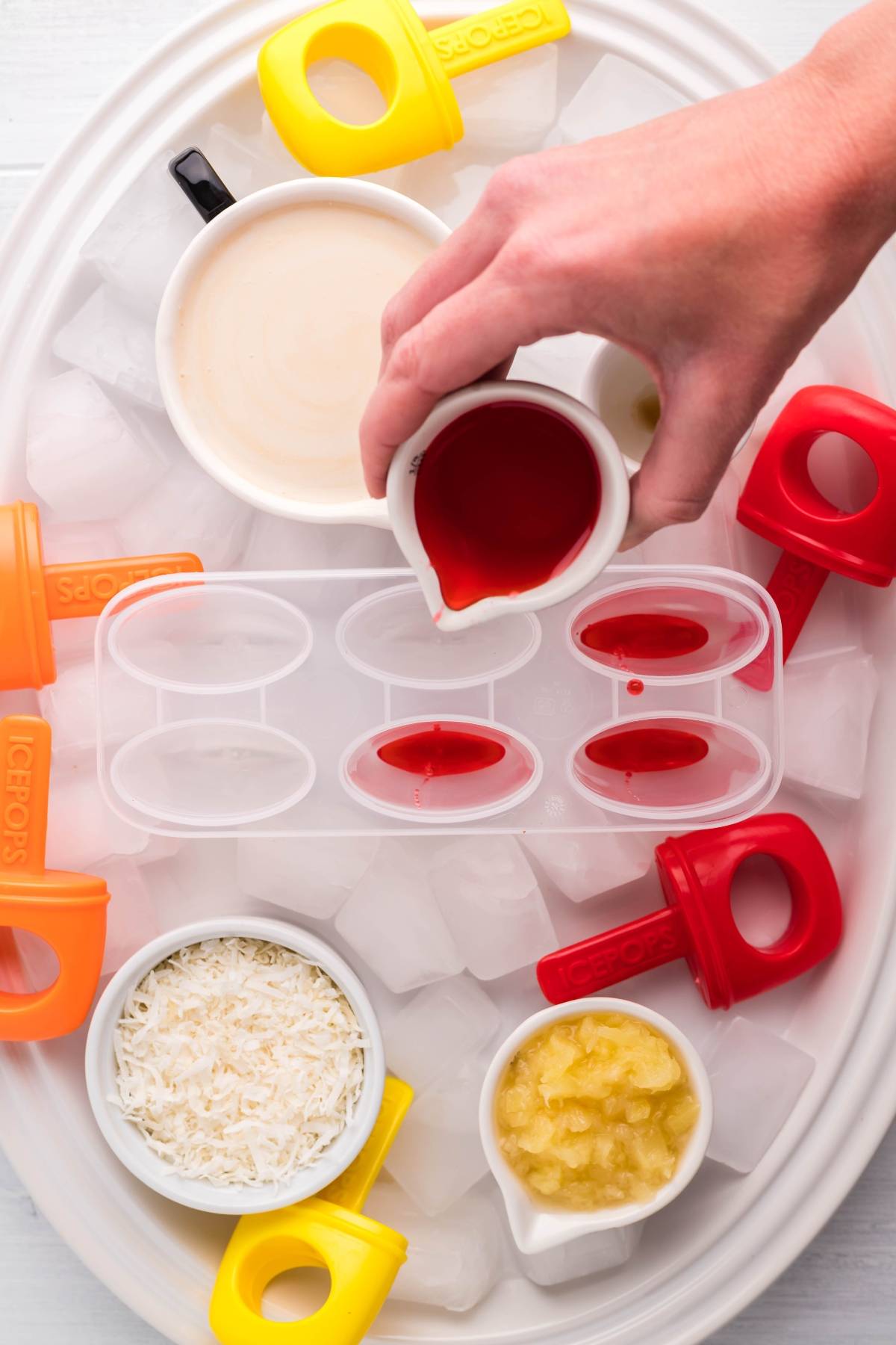 A hand pours red liquid into popsicle molds, surrounded by ice, bowls of coconut, pineapple, and pop molds—perfect for making a layered popsicle recipe or delicious sugar free popsicles.