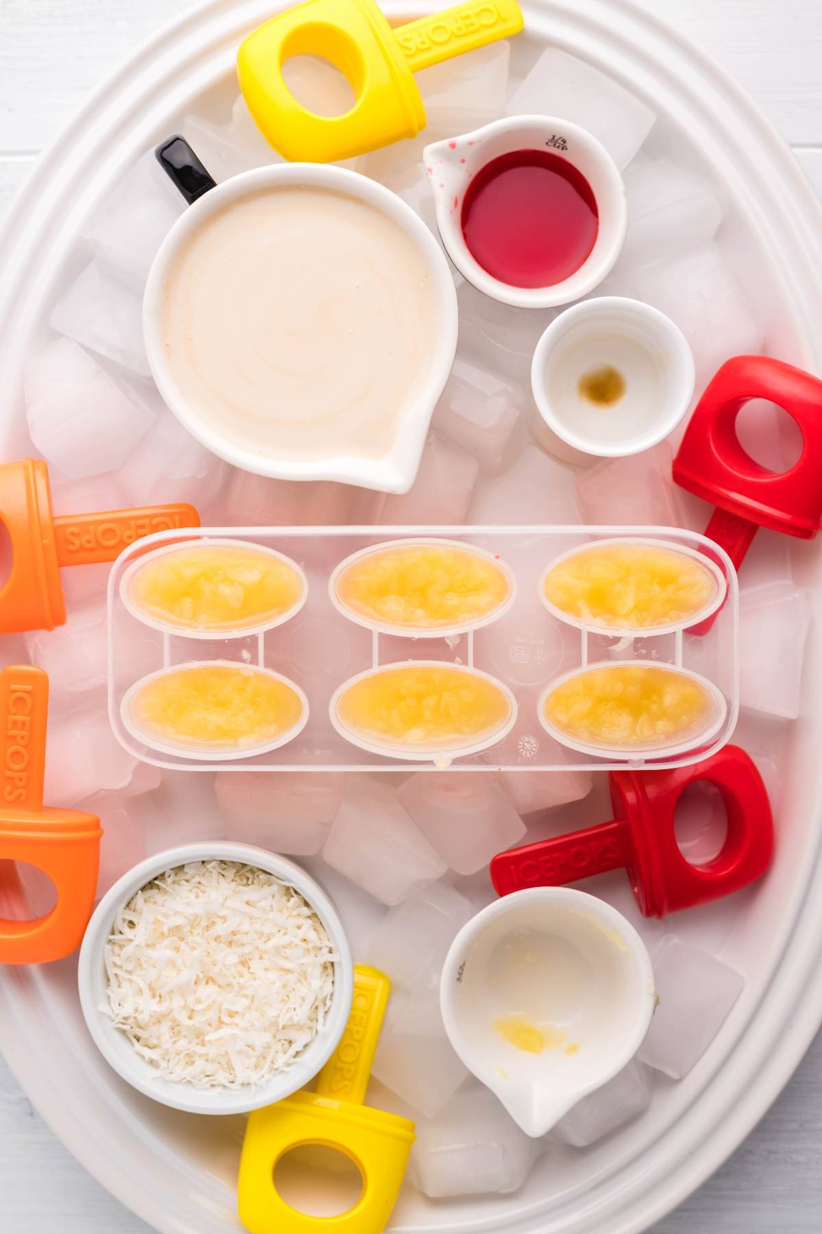 Oval tray with ice, coconut popsicle molds, shredded coconut, creamy liquid, red syrup, and chopped pineapple—perfect for making Sugar Free Popsicles or delicious Tropical Sunrise Popsicles.