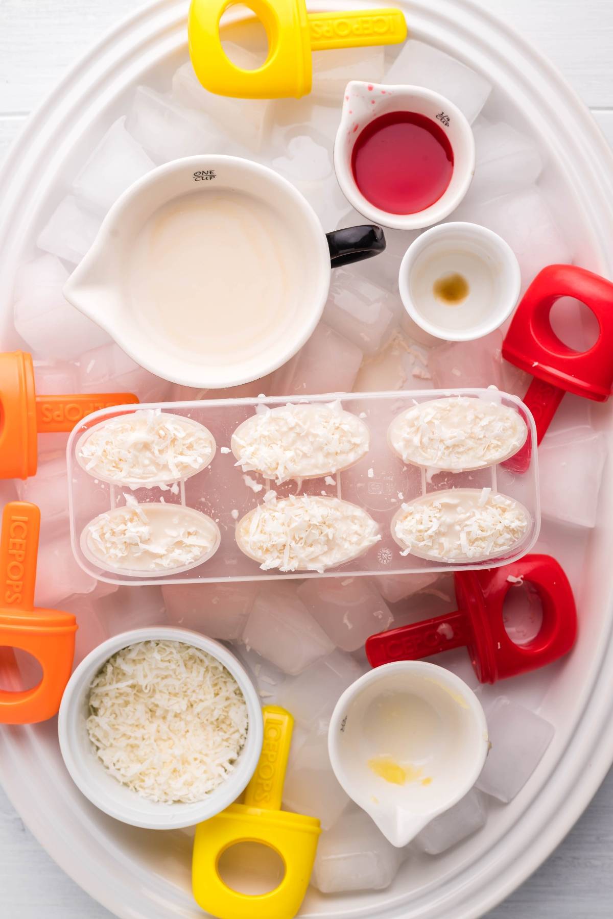 Tropical Sunrise Popsicles mold filled with creamy mixture, shredded coconut, and colorful handles rests on a tray of ice alongside small bowls—perfect for making delicious coconut ice pops.
