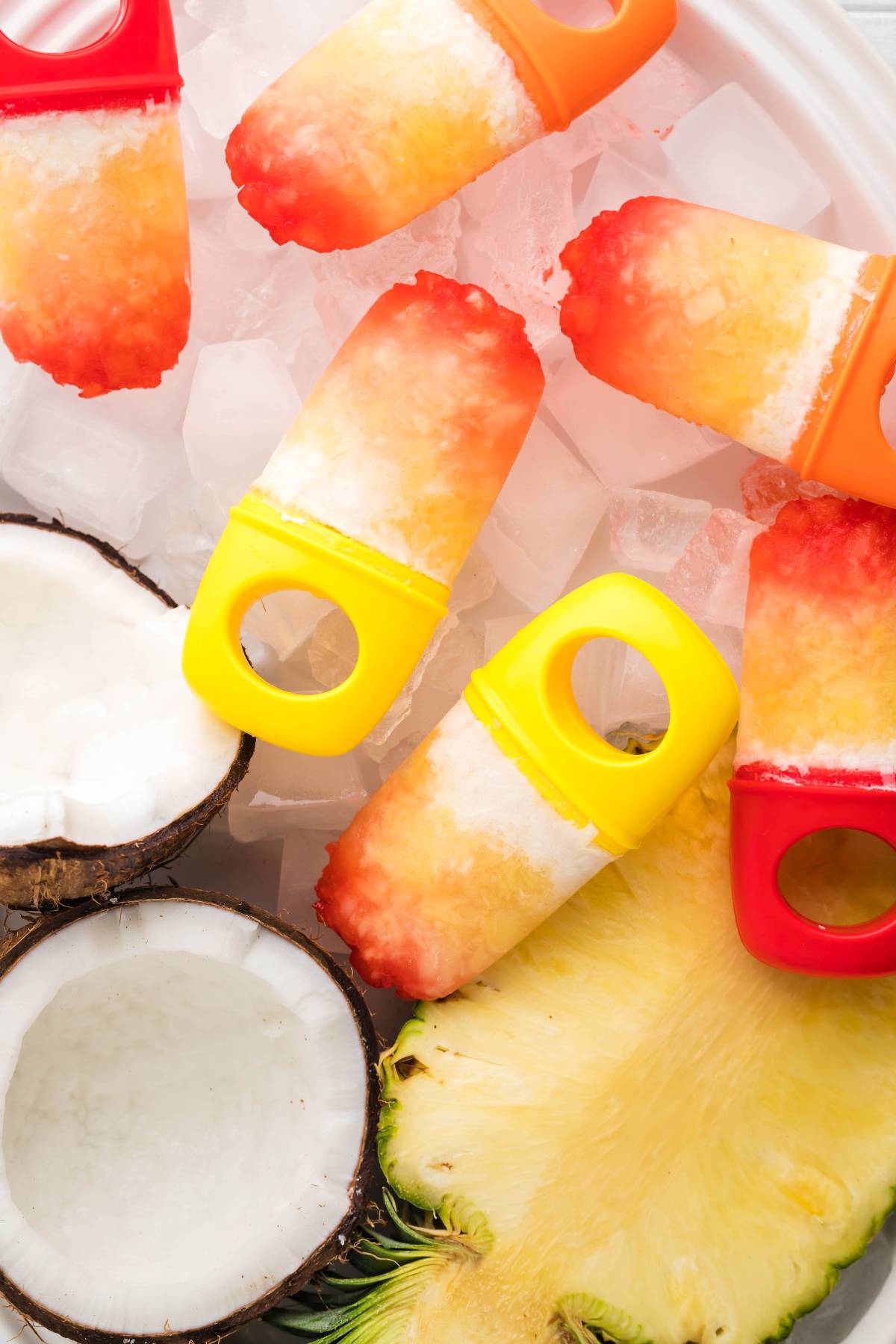 Colorful Coconut ice pops rest on ice, surrounded by coconut halves and pineapple slices for a refreshing tropical treat.