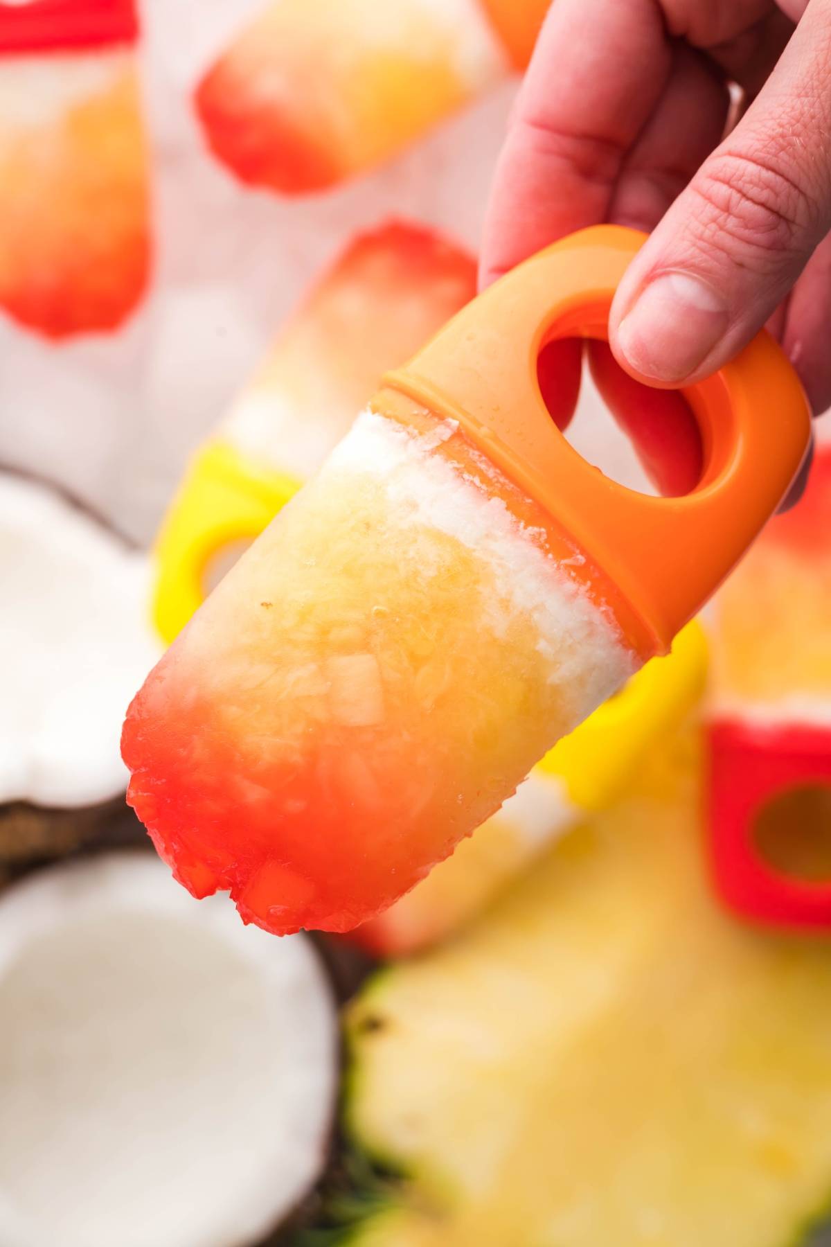 A hand holding a colorful, homemade Tropical Sunrise Popsicle in a bright orange mold.