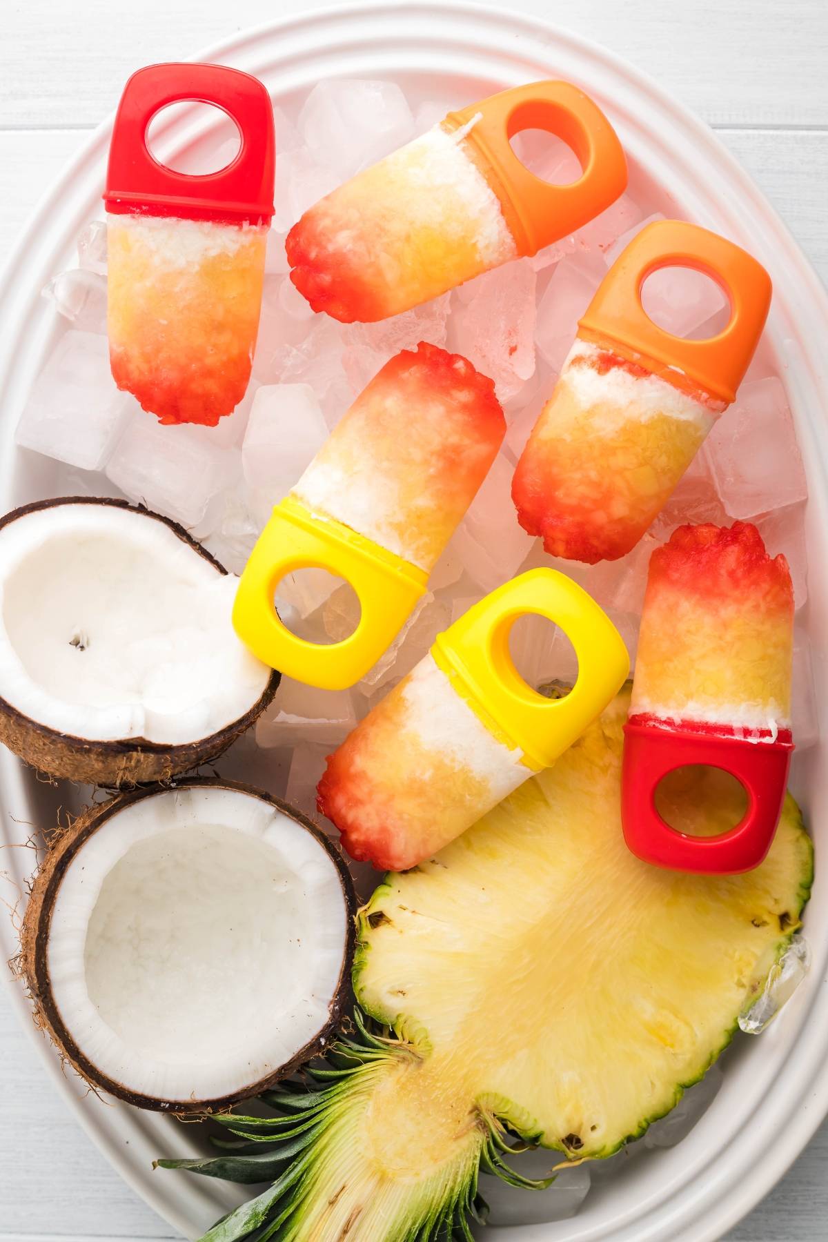 Tropical Sunrise Popsicles shaped like rings rest on ice, with sliced coconut and pineapple, creating vibrant Coconut ice pops on a white platter.