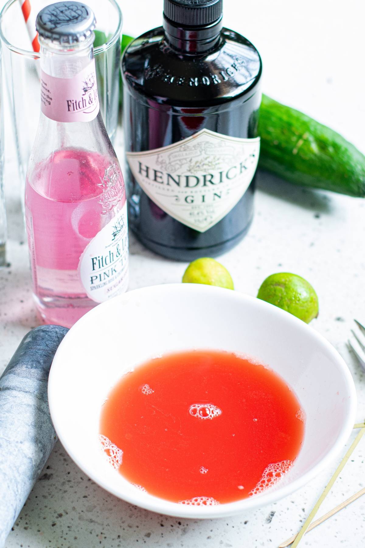 A bowl of red liquid with gin, pink tonic, cut cucumber ribbons, and limes on a white countertop—perfect as a refreshing summer BBQ cocktail or for trying a new summer gin and tonic recipe.