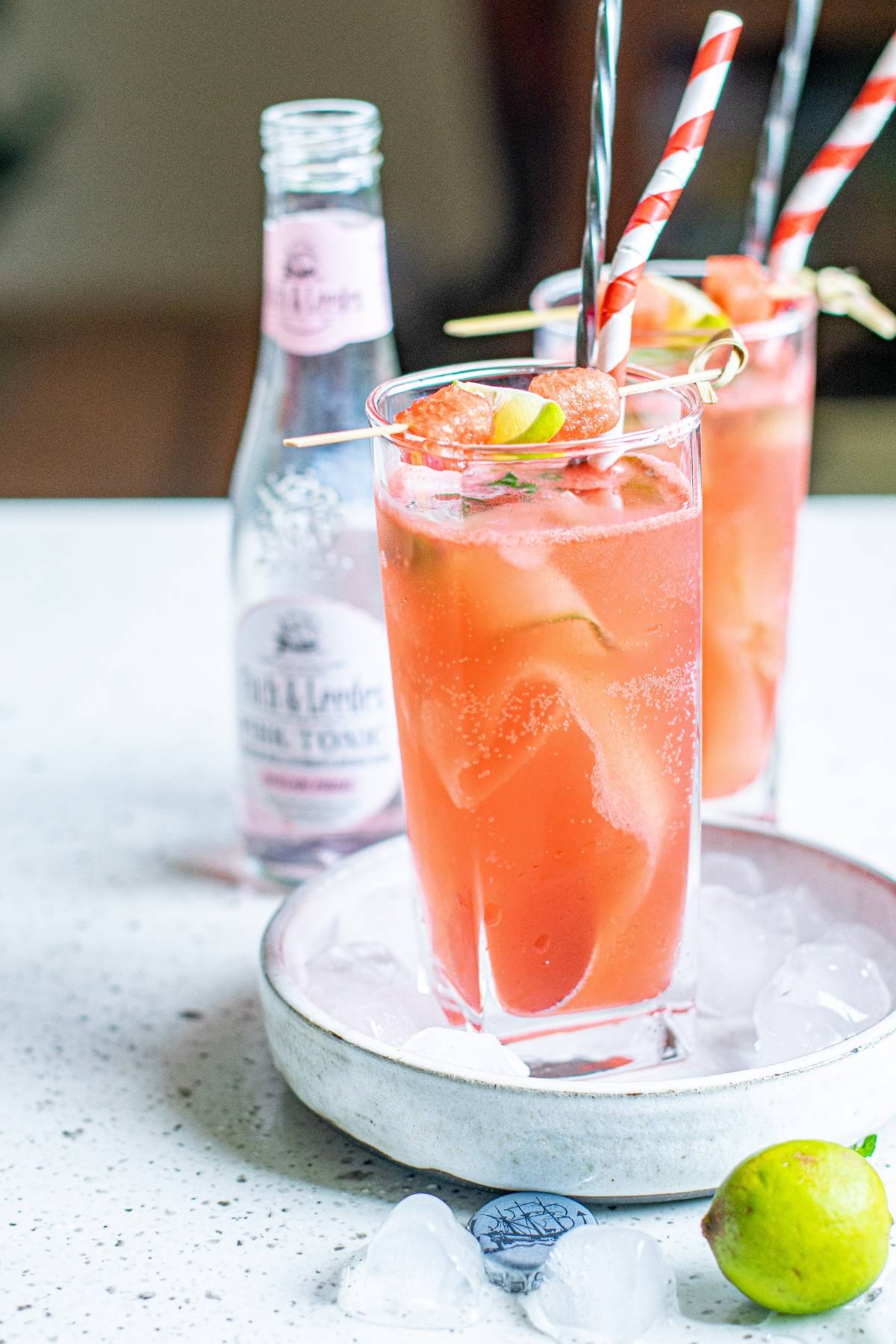 Two tall glasses of pink Watermelon Gin Tonic with straws and lime, garnished with grapefruit and cut cucumber ribbons, sit beside a tonic water bottle—a refreshing summer BBQ cocktail.