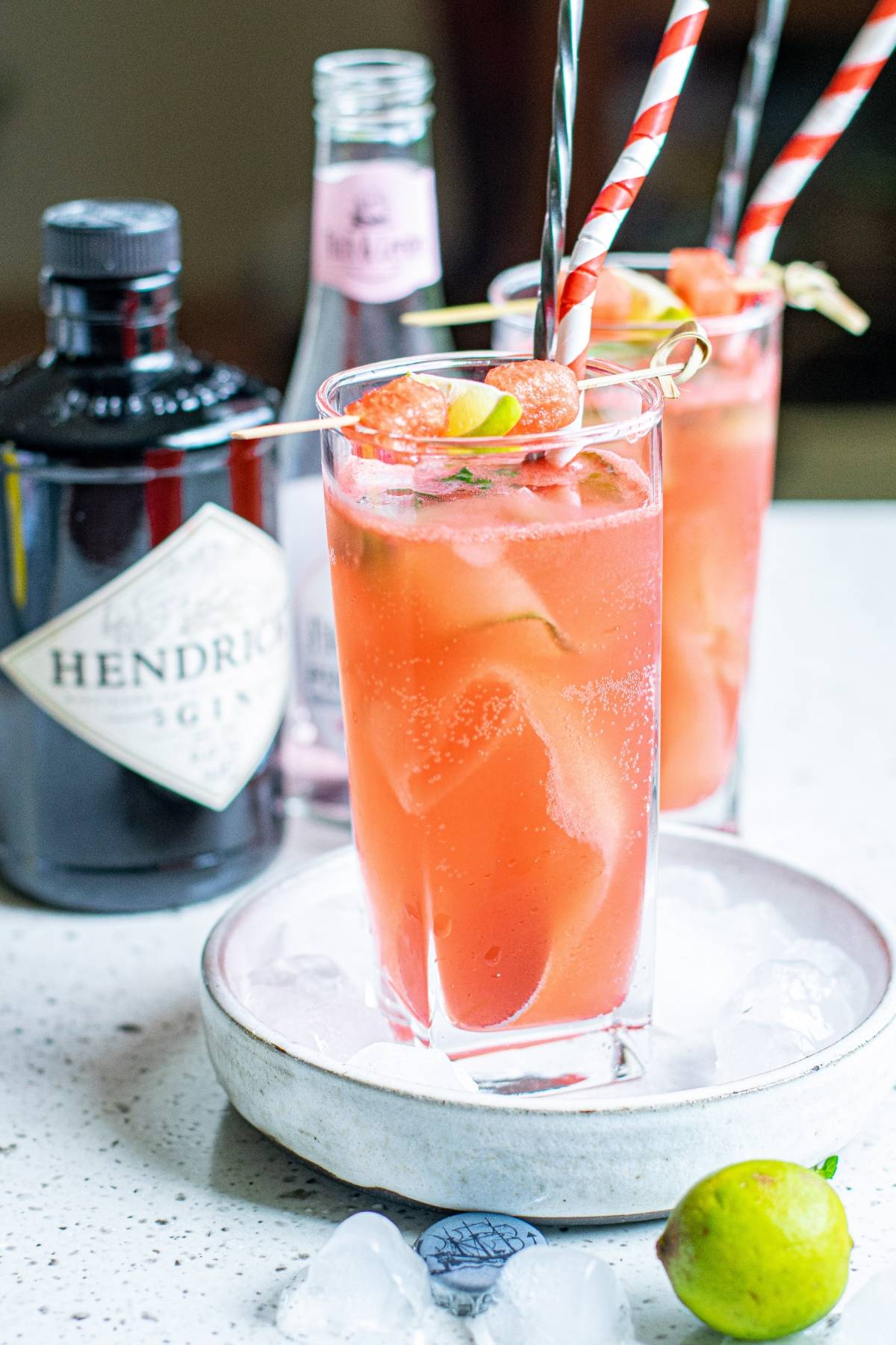 Two pink gin cocktails with striped straws, surrounded by gin bottle, tonic, lime, cut cucumber ribbons, and ice on a white tray—perfect for trying a refreshing Summer gin and tonic recipe.