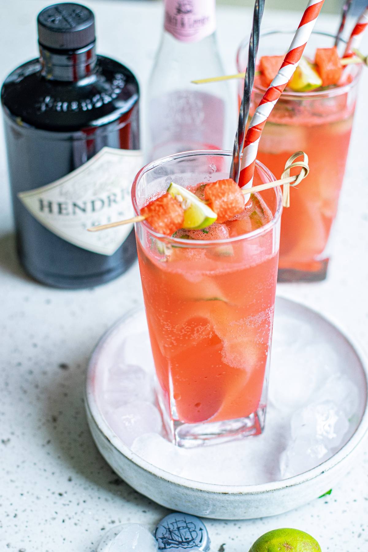 A tall pink cocktail with ice, lime, cut cucumber ribbons, and grapefruit garnish sits beside a Hendrick’s gin bottle and tonic water—your refreshing summer BBQ cocktail.