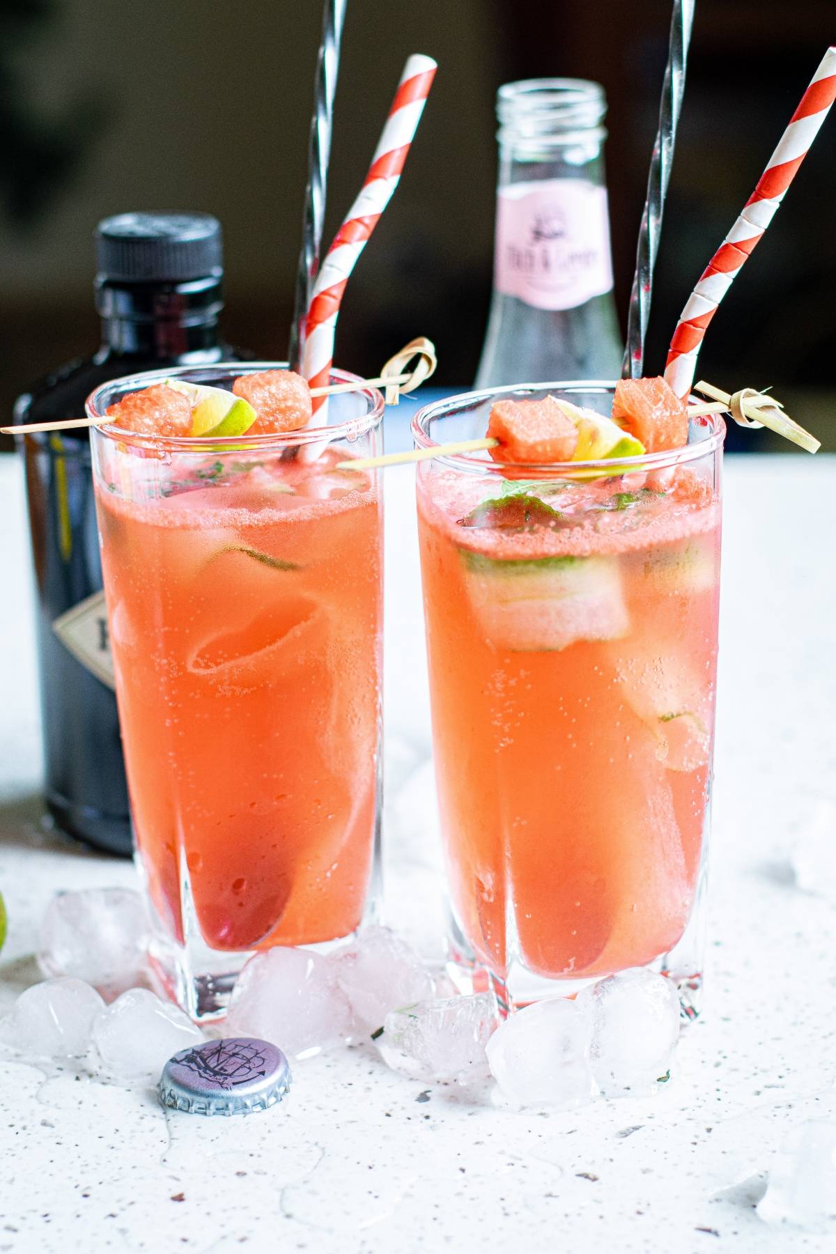Two tall glasses of pink cocktail with ice, lime slices, cut cucumber ribbons, and paper straws, surrounded by ice cubes and bottles—a refreshing nod to a watermelon cucumber drink.