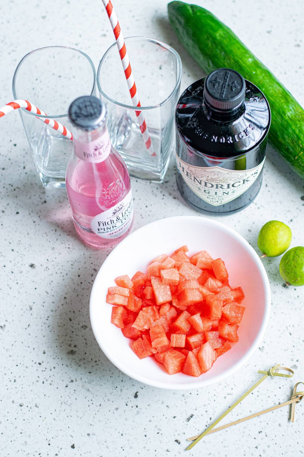 Bowl of diced watermelon, gin bottle, tonic, cut cucumber ribbons, limes, glasses, and straws on a countertop—perfect for crafting a refreshing Watermelon Gin Tonic or your new favorite summer gin and tonic recipe.