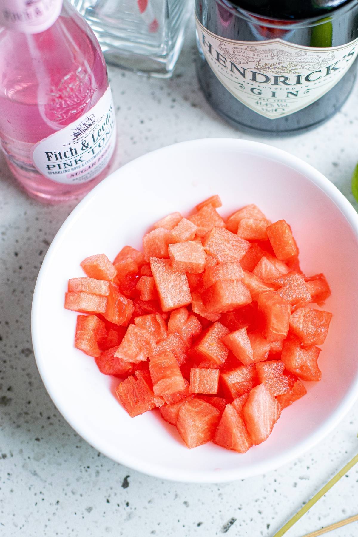 A bowl of diced watermelon sits beside a bottle of gin, a pink tonic water bottle, and some cut cucumber ribbons—perfect ingredients for a refreshing Watermelon Gin Tonic.