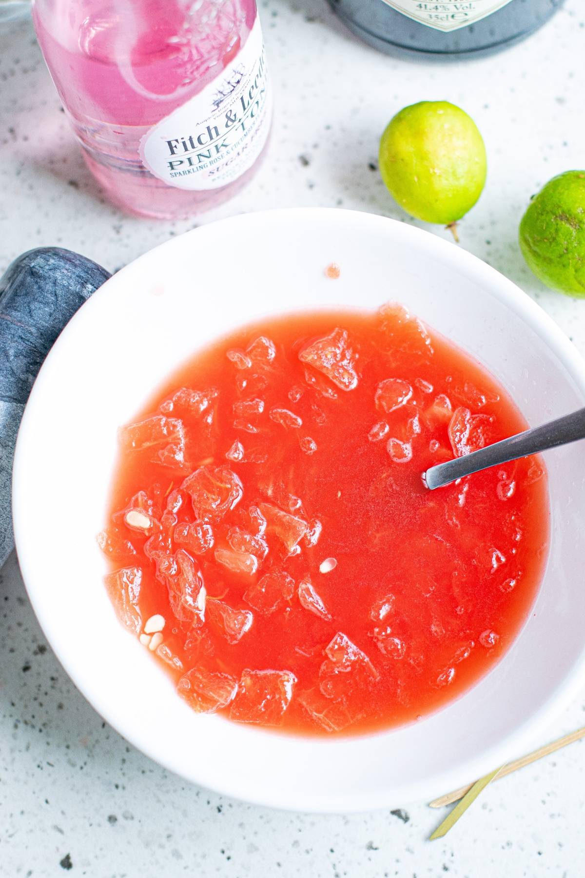 A white bowl with crushed ice and red liquid, next to limes, cut cucumber ribbons, and a pink tonic bottle on a countertop—a refreshing summer BBQ cocktail scene.
