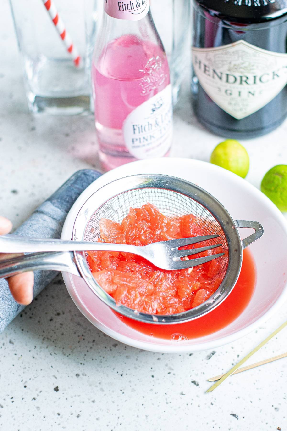 A fork presses grapefruit pulp in a strainer over a bowl, with bottles, limes, and cut cucumber ribbons ready for a refreshing summer BBQ cocktail like a Watermelon Gin Tonic.
