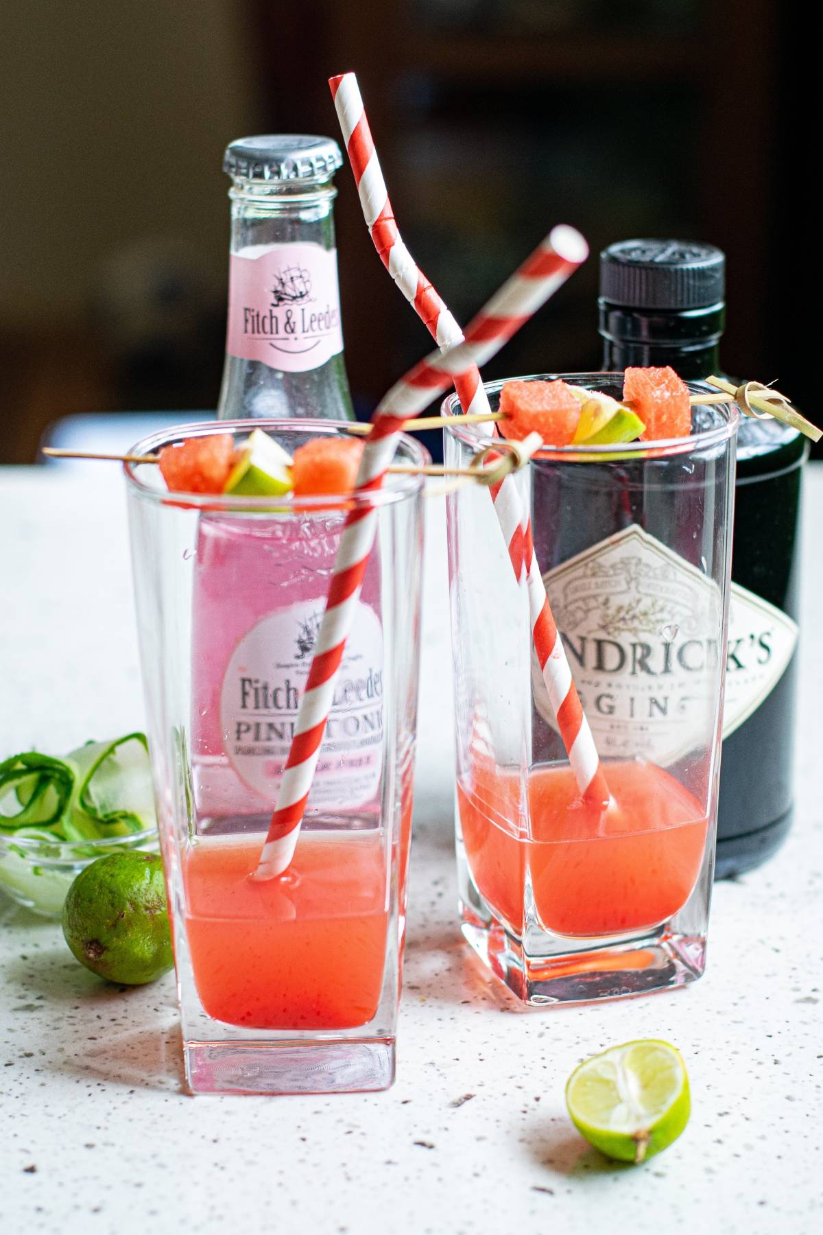 Two glasses of pink gin cocktails with striped straws, garnished with grapefruit and lime, sit beside drink bottles—perfect for trying a refreshing summer gin and tonic recipe like a Watermelon Gin Tonic.