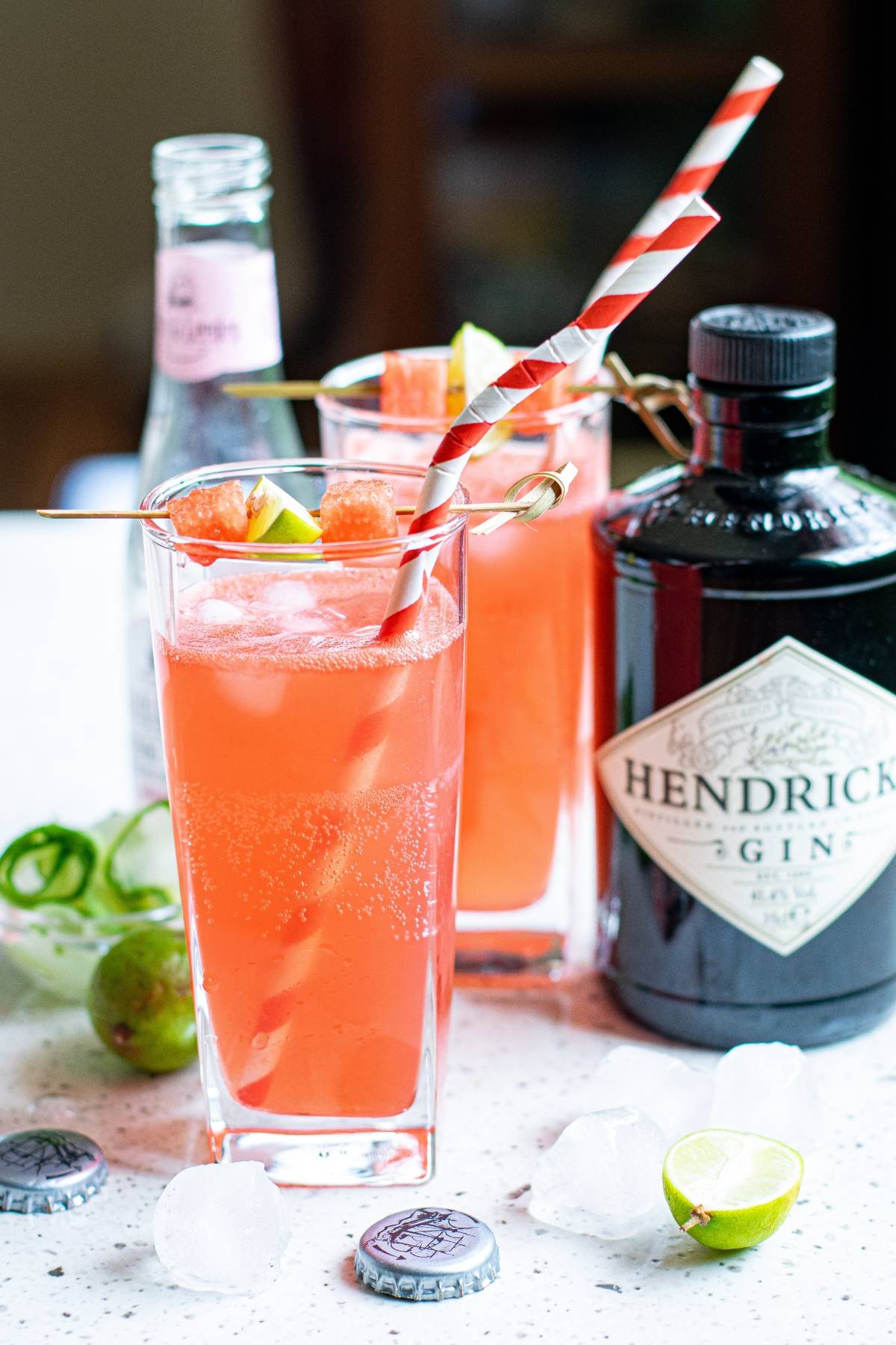 Two pink gin cocktails with striped straws, garnished with lime and cut cucumber ribbons, sit beside a Hendrick's gin bottle and ice.