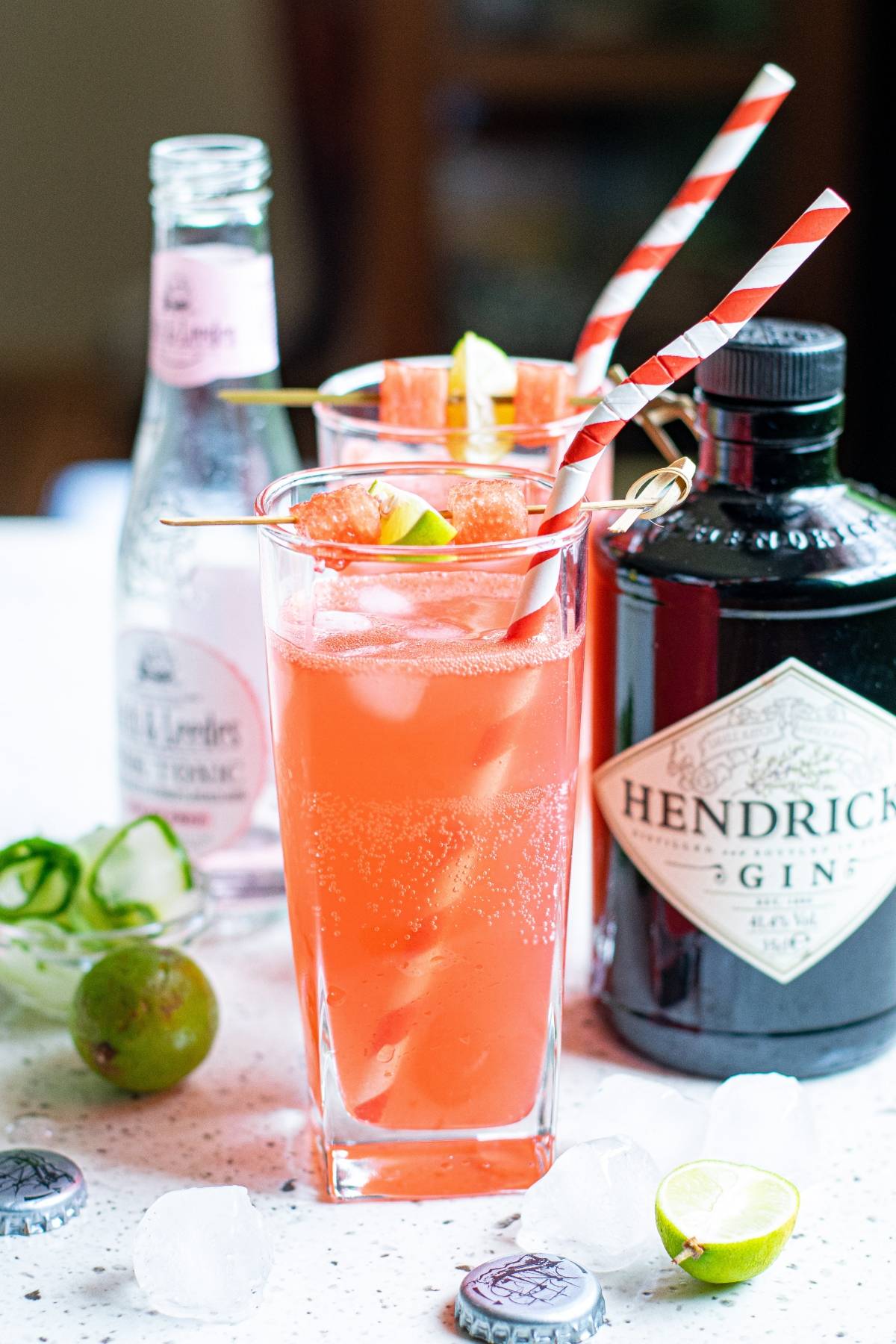 Two pink gin cocktails with lime, striped straws, tonic bottle, gin bottle, ice, and limes on a table—the perfect refreshing summer BBQ cocktail for any gathering.