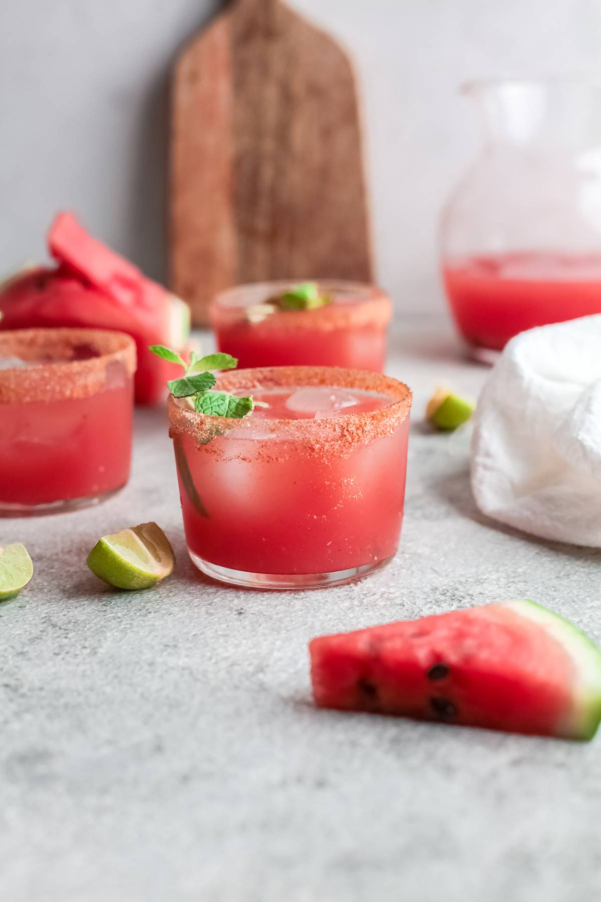 A glass of cocktail made with watermelon and a chili rim, garnished with mint, surrounded by lime wedges and watermelon slices—an easy summer cocktail recipe perfect for refreshing moments.