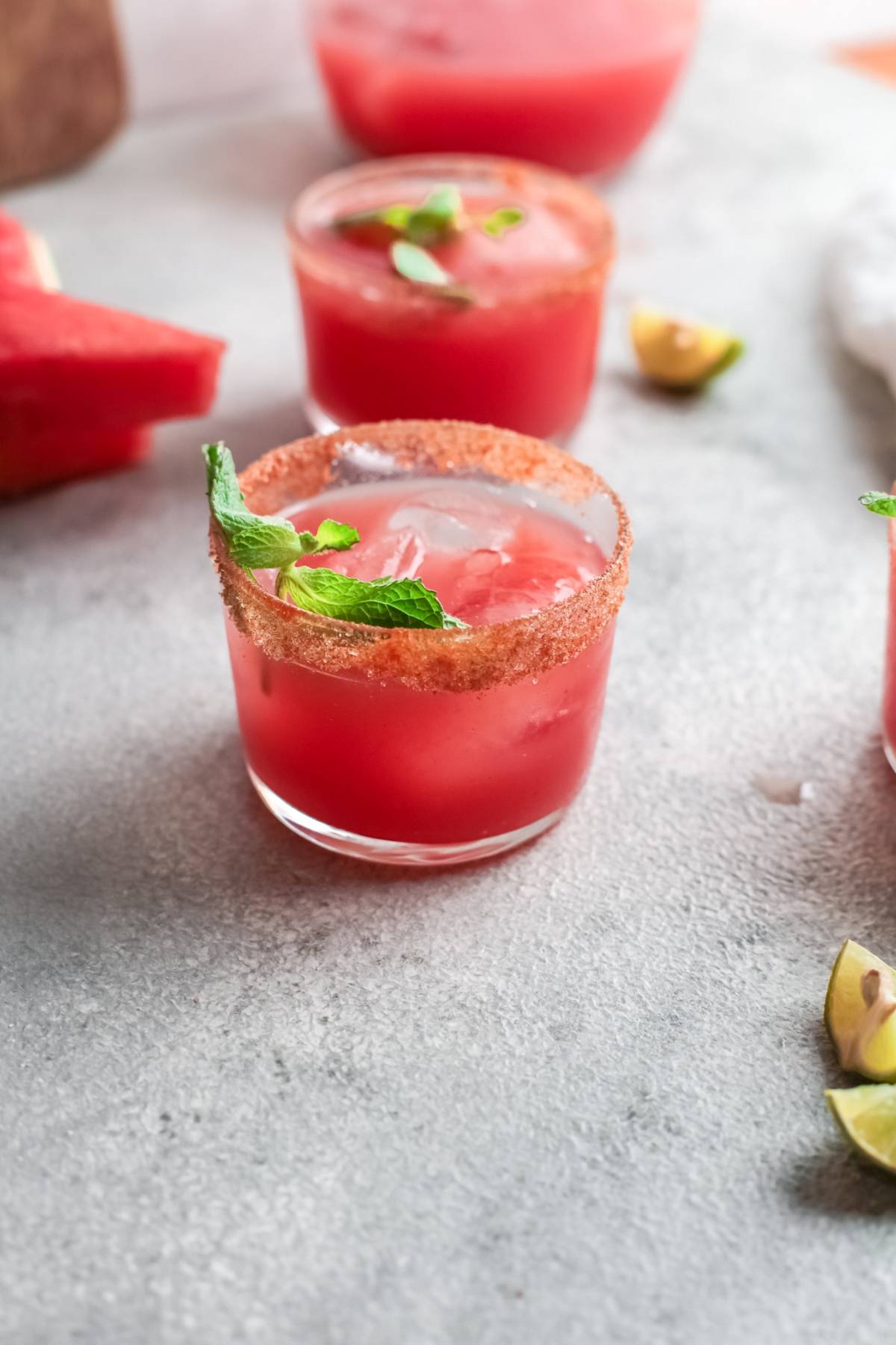 A glass of pink Watermelon Paloma with a chili rim and mint garnish on a gray surface, made with watermelon juice for a tangy and sweet paloma twist.