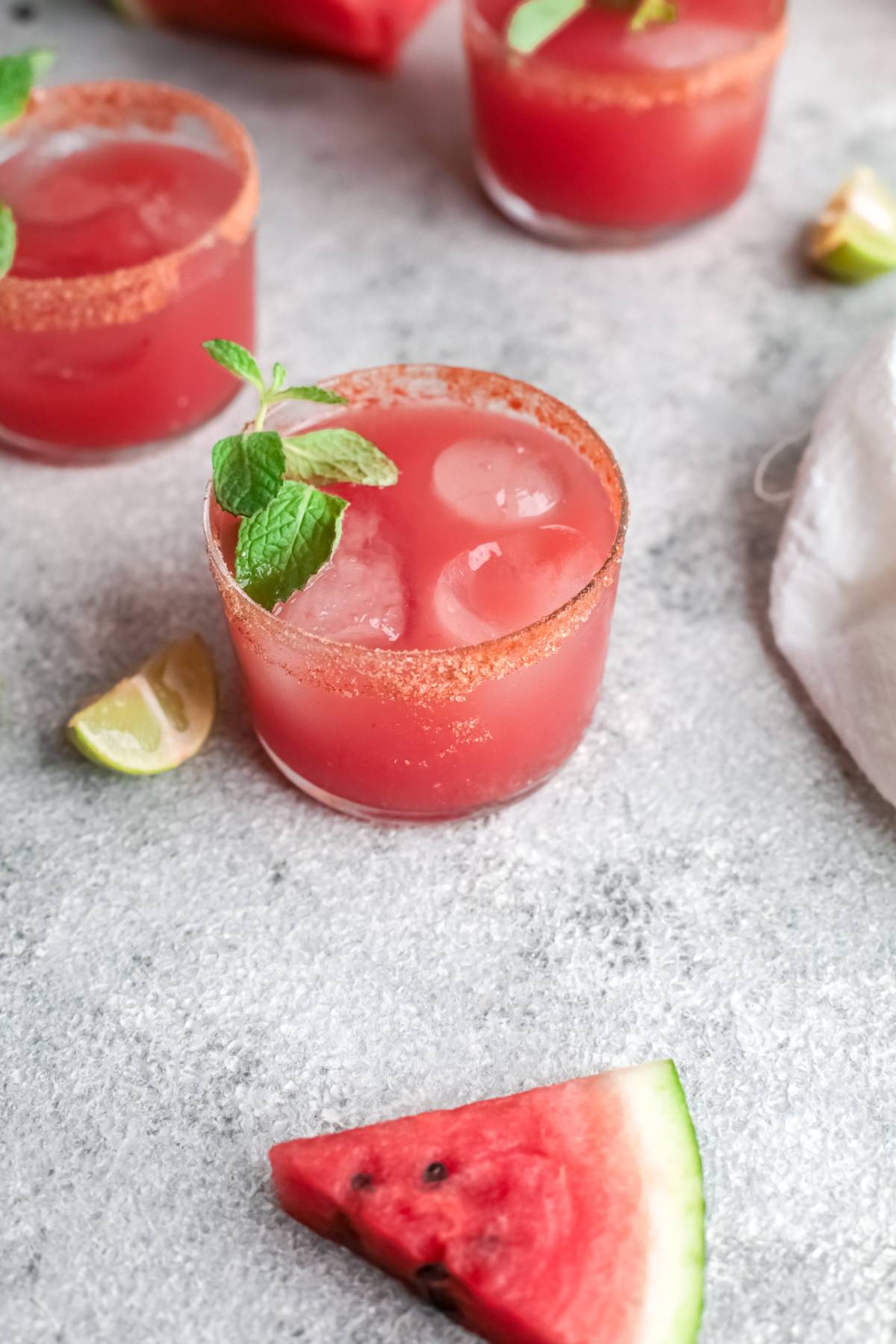 A glass of Watermelon Paloma with ice, mint garnish, and chili rim sits next to a lime wedge and watermelon slice—this tangy and sweet paloma is made with watermelon juice for a refreshing twist.