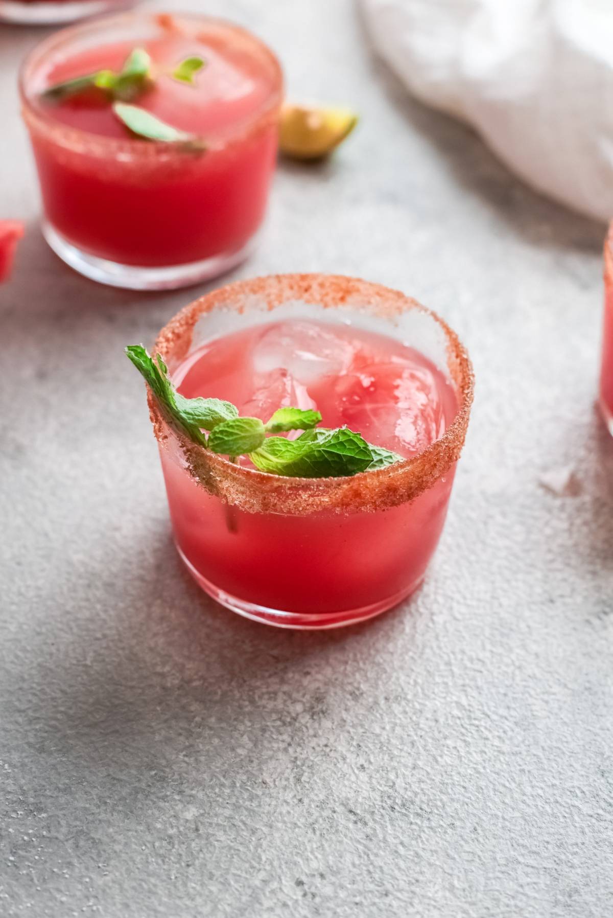 A pink cocktail made with watermelon juice, ice, mint garnish, and a spiced rim in a glass on a gray surface.