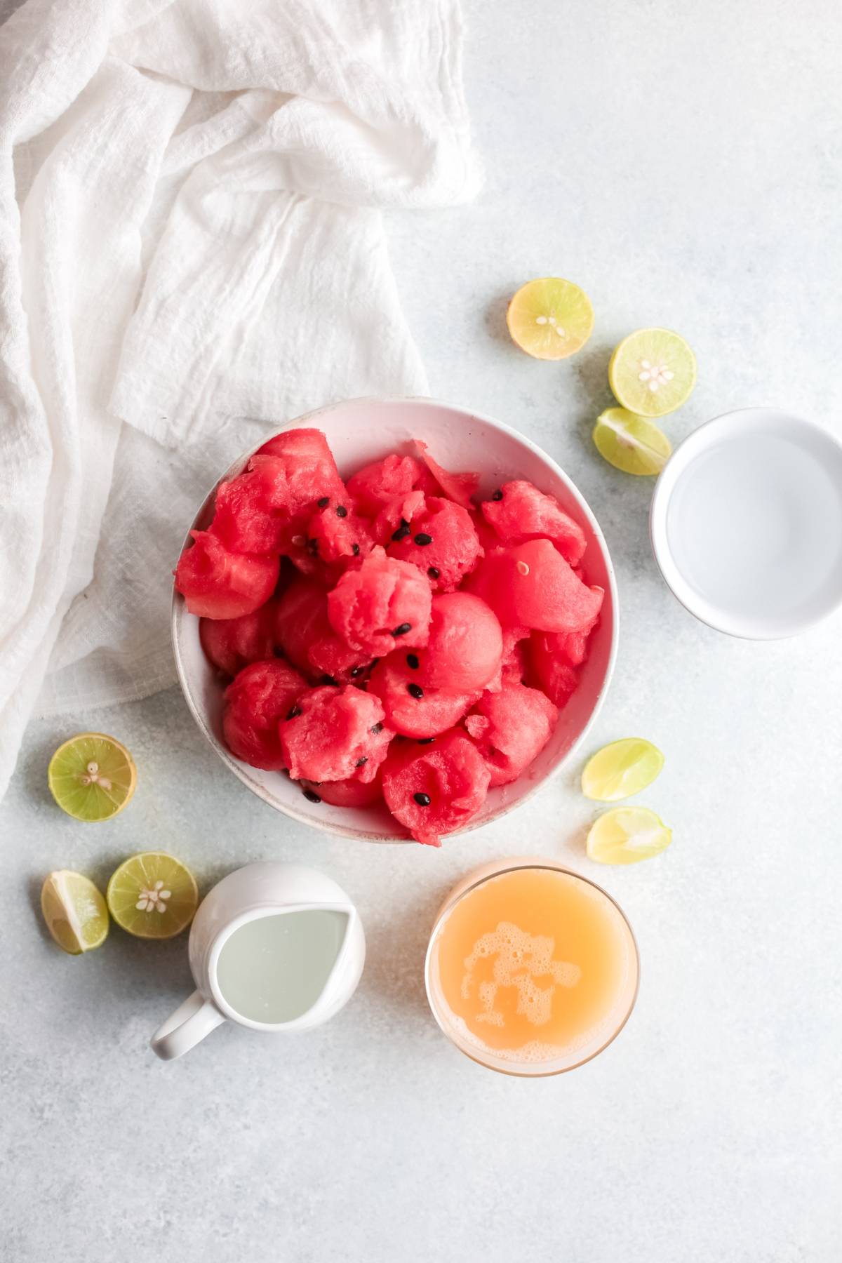Bowl of watermelon balls, sliced limes, a glass of juice, and a small white pitcher on a light surface—perfect for an easy summer cocktail recipe like a refreshing Watermelon Paloma.