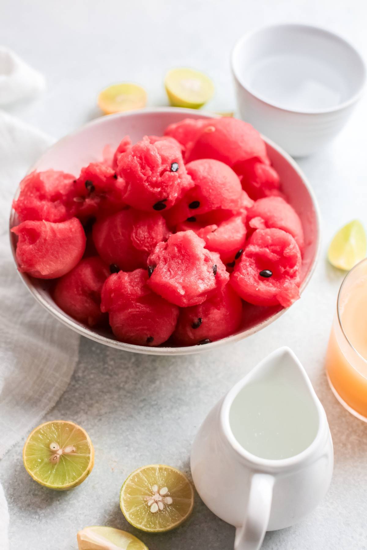 A bowl of watermelon balls with lime halves is paired with a pitcher of milk, plus glasses of water and juice—perfect inspiration for an easy summer cocktail recipe or a tangy and sweet paloma cocktail made with watermelon.