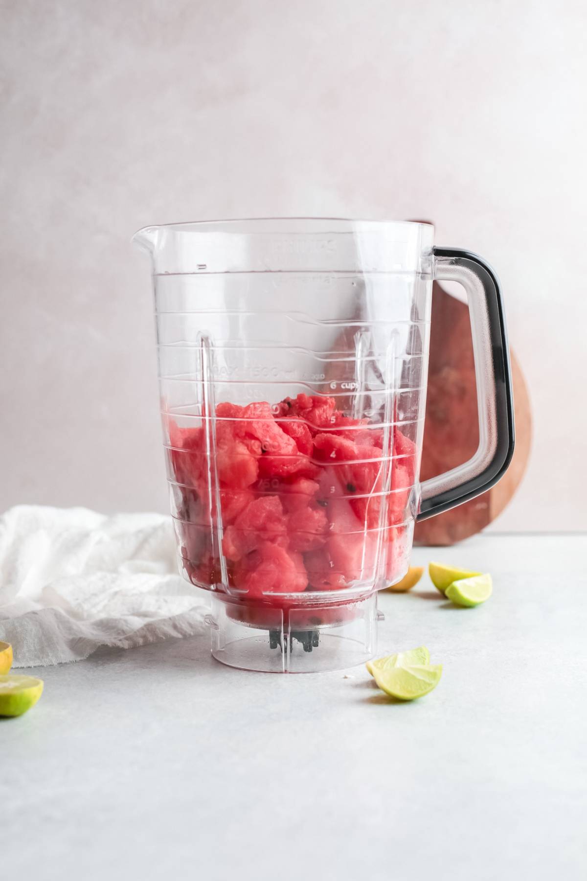 A blender pitcher filled with chopped watermelon sits on a counter with lime wedges nearby, ready to be transformed into a tangy and sweet Watermelon Paloma cocktail.