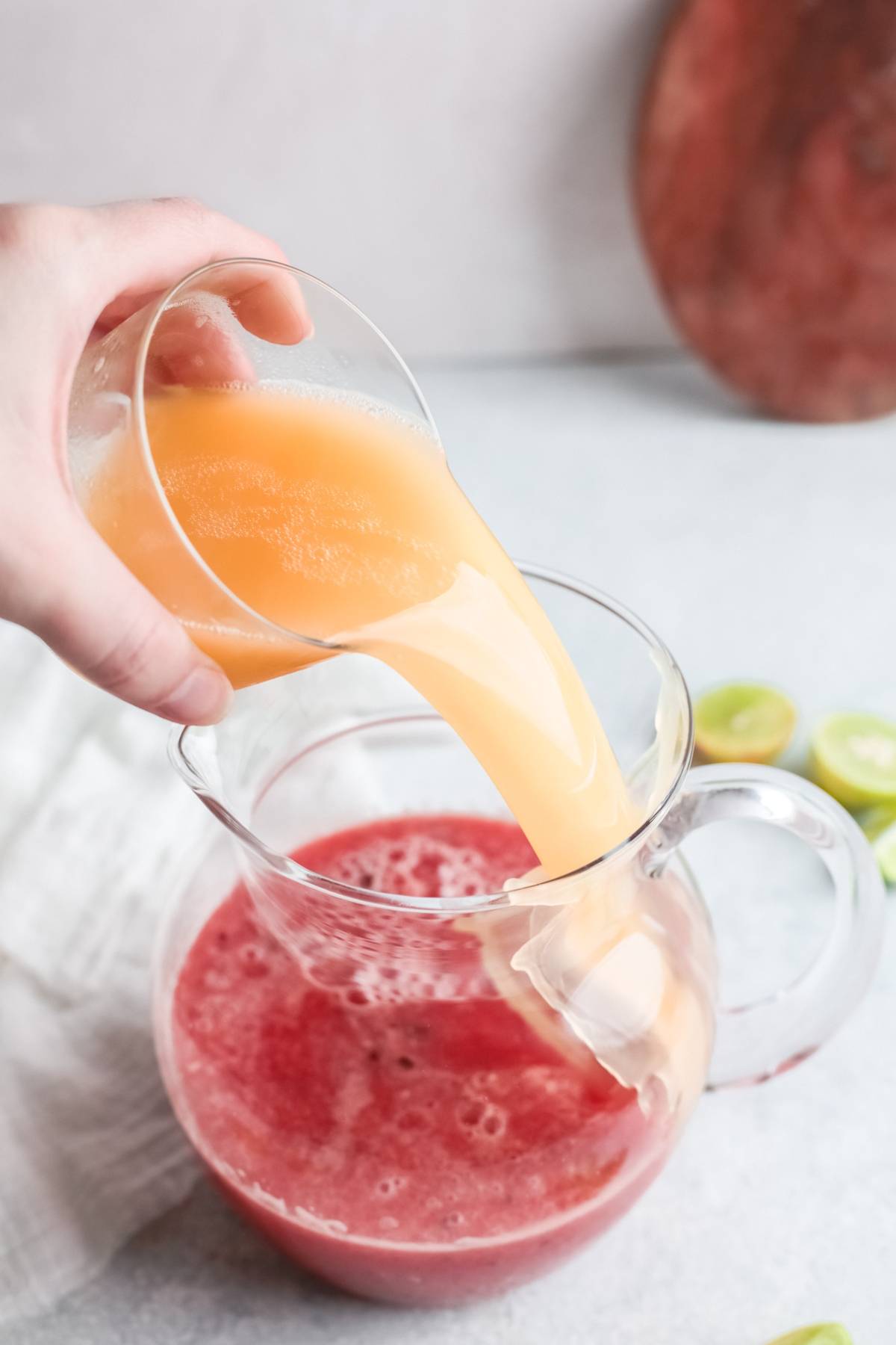 A hand pours orange juice from a glass into a clear pitcher of red juice on a light surface—a refreshing cocktail made with watermelon for a tangy and sweet paloma twist.
