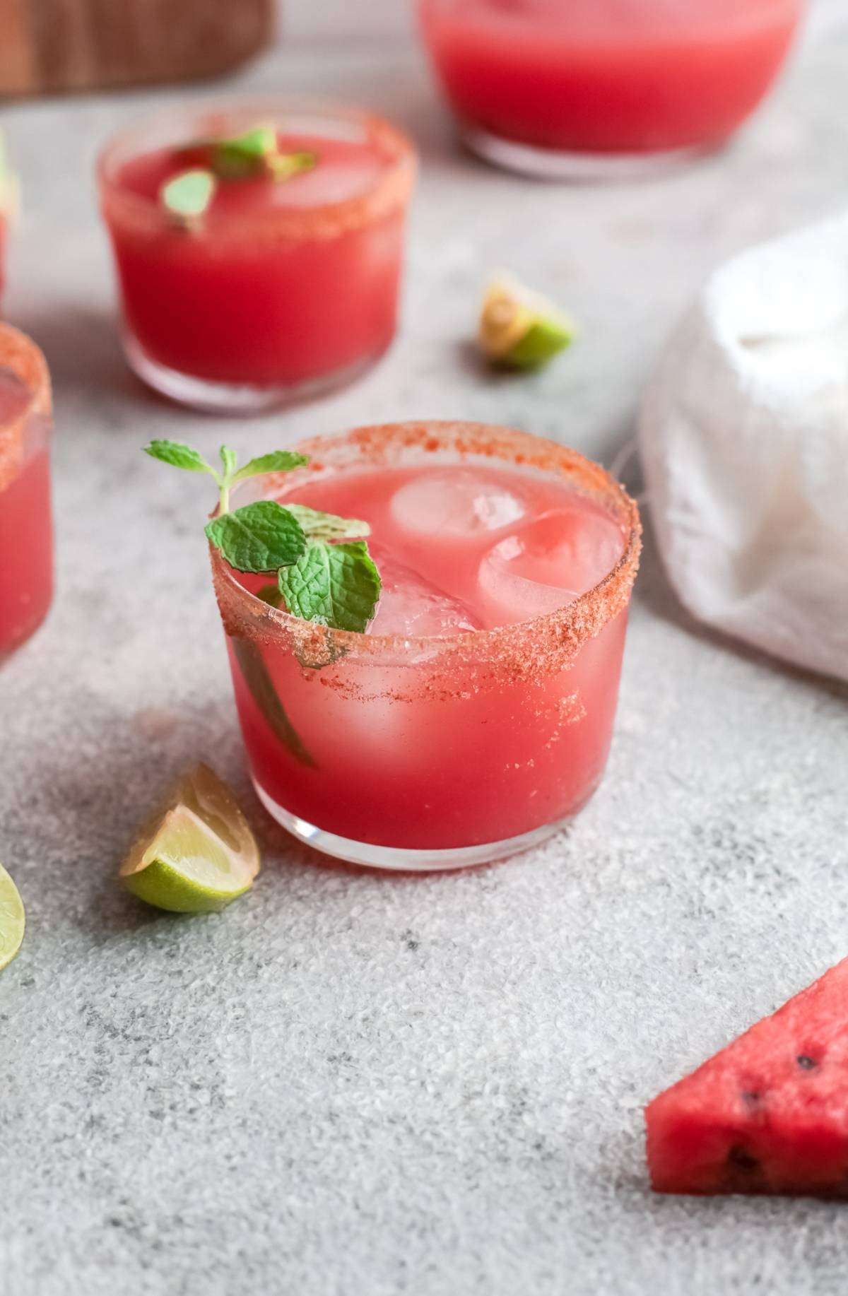 A glass of pink Watermelon Paloma with ice, mint, and chili rim, surrounded by lime wedges and a watermelon slice. An easy summer cocktail recipe made with watermelon juice for a refreshing twist.