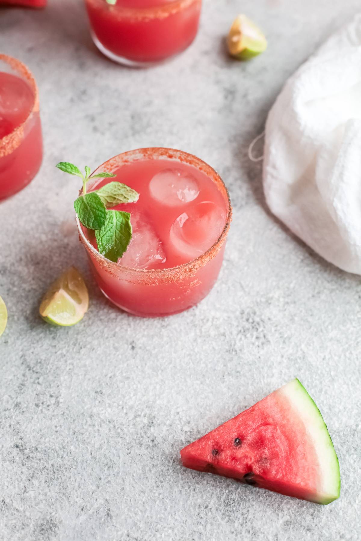 A glass of Watermelon Paloma with ice, mint garnish, and a chili rim sits next to lime wedges and a watermelon slice—an easy summer cocktail recipe perfect for warm days.
