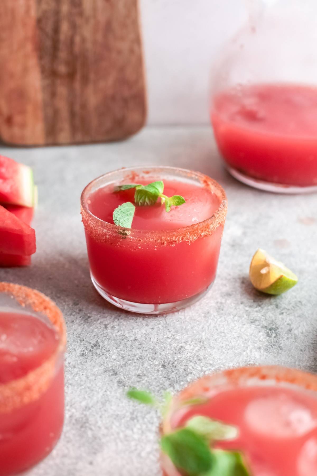 A glass of tangy and sweet paloma made with watermelon juice and ice, garnished with mint and a spiced rim, sits on a gray surface.