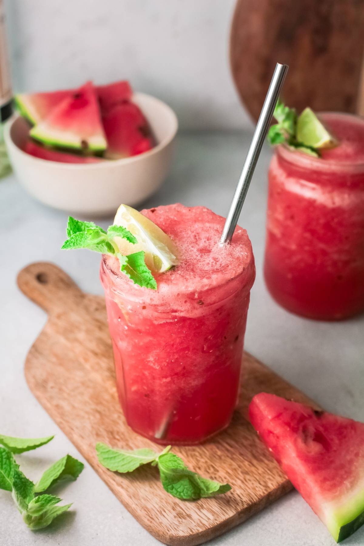 A frosty Watermelon White Wine Slushie in a jar with lime, mint, and a metal straw sits on a wooden board—the perfect frozen watermelon cocktail for summer.