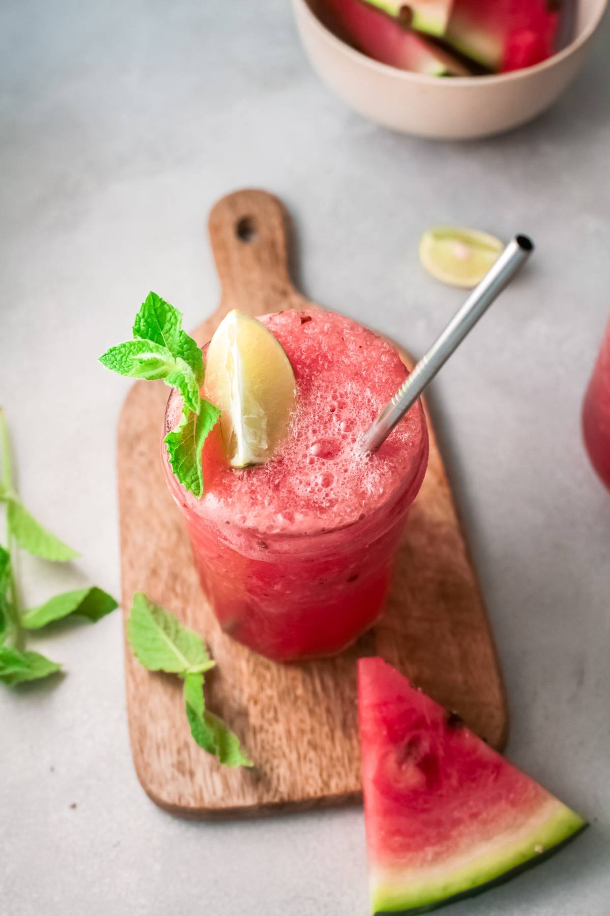 A glass of Frozen watermelon cocktail with mint, lemon, and a straw on a wooden board, surrounded by fresh watermelon slices.
