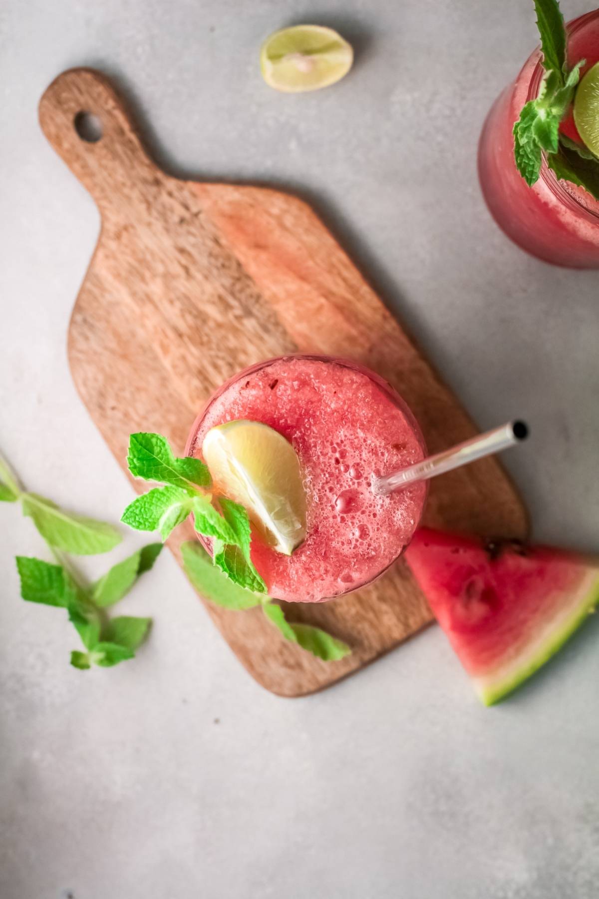 Top view of a Watermelon White Wine Slushie with mint and lime on a wooden board, next to a watermelon slice.