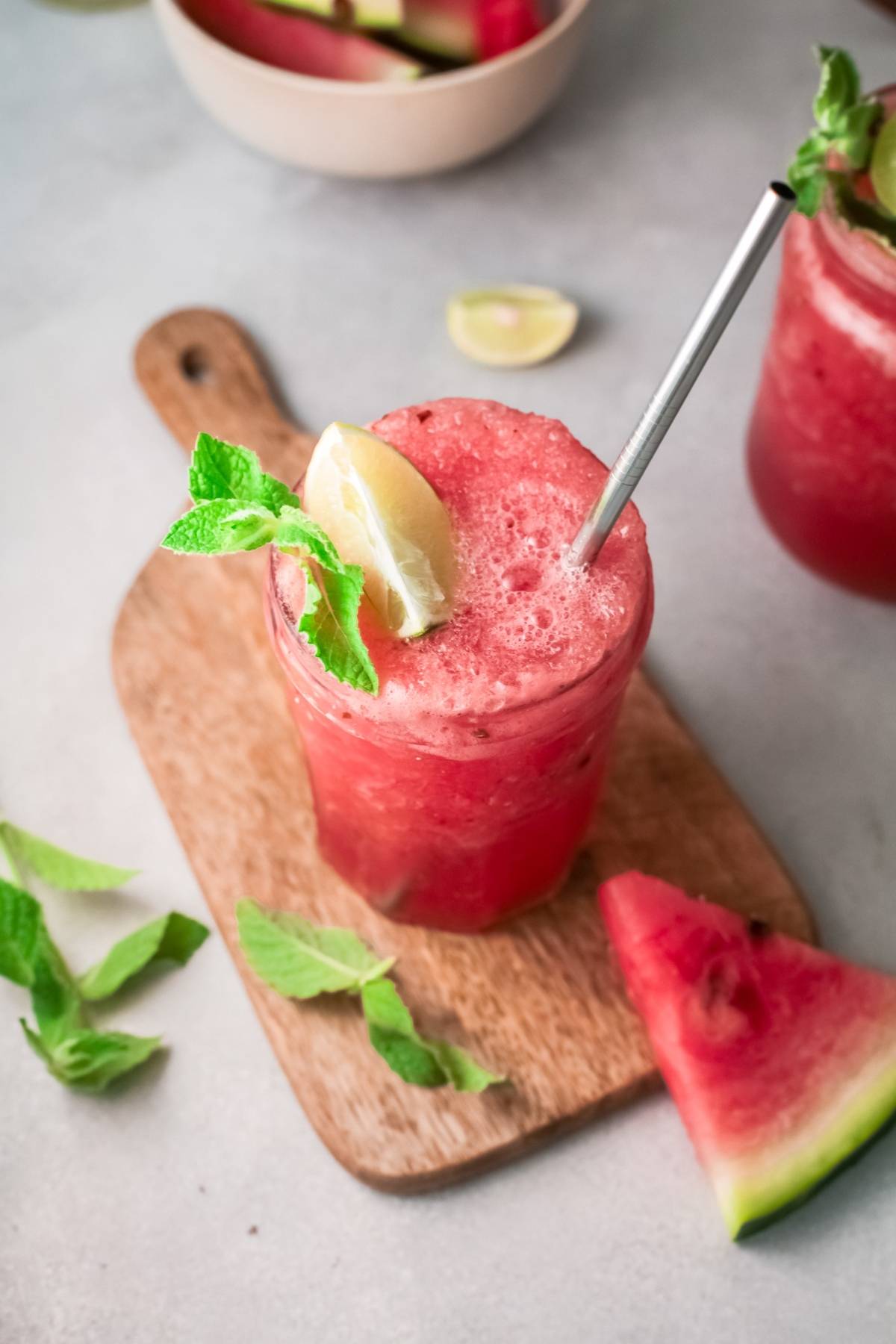 A refreshing alcoholic slushie drink recipe: a watermelon drink with mint, lime, and a straw on a wooden board, garnished with a watermelon slice. Perfect as a summer recipe using watermelon and wine.