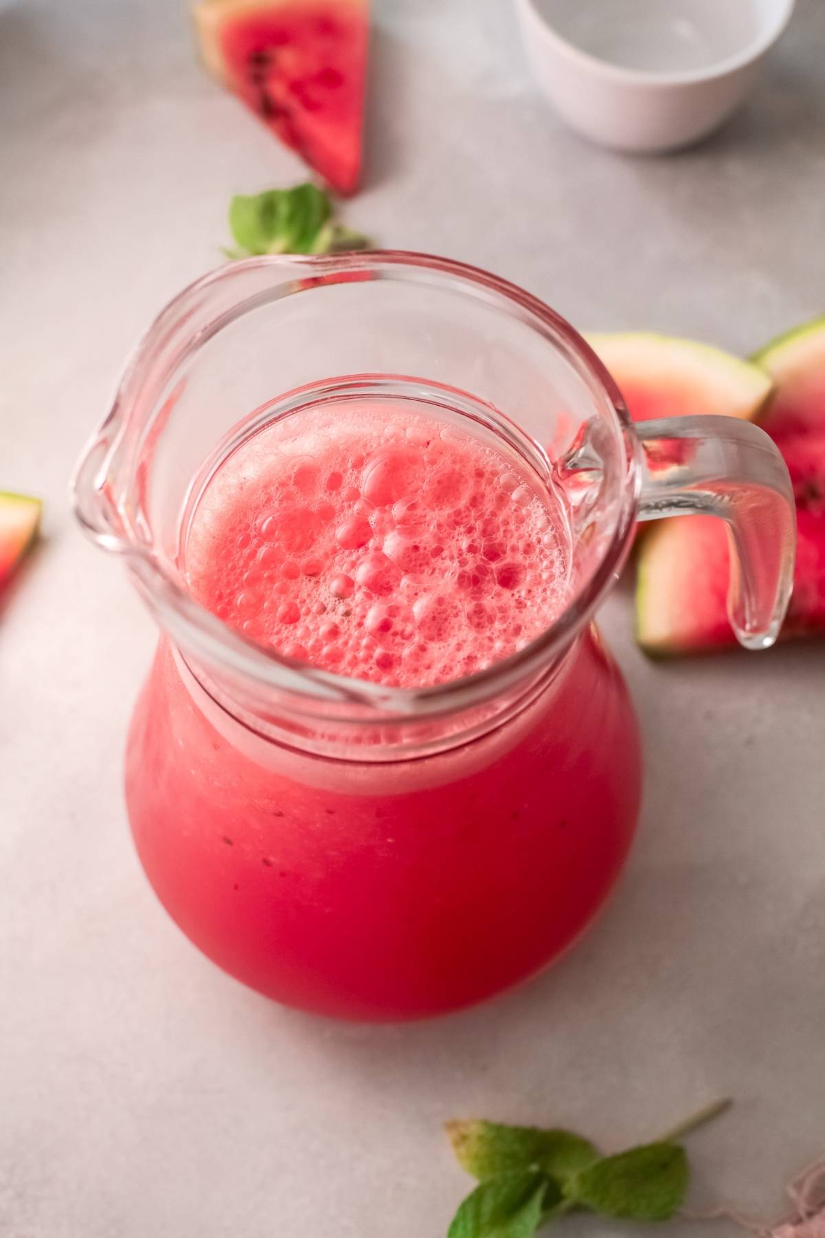 A glass pitcher filled with frothy pink watermelon juice, surrounded by watermelon slices and mint leaves—perfect for a refreshing Frozen Watermelon Cocktail or a tasty Recipe using watermelon and wine.