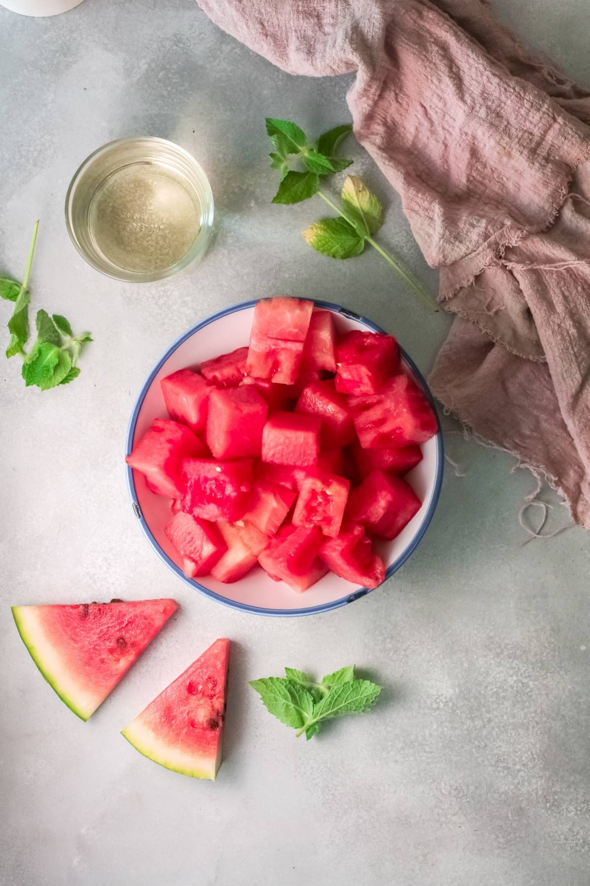 A bowl of cubed watermelon, two watermelon slices, mint leaves, a glass of water, and a pink cloth on a table—perfect inspiration for your next frozen watermelon cocktail or refreshing alcoholic slushie drink recipe.