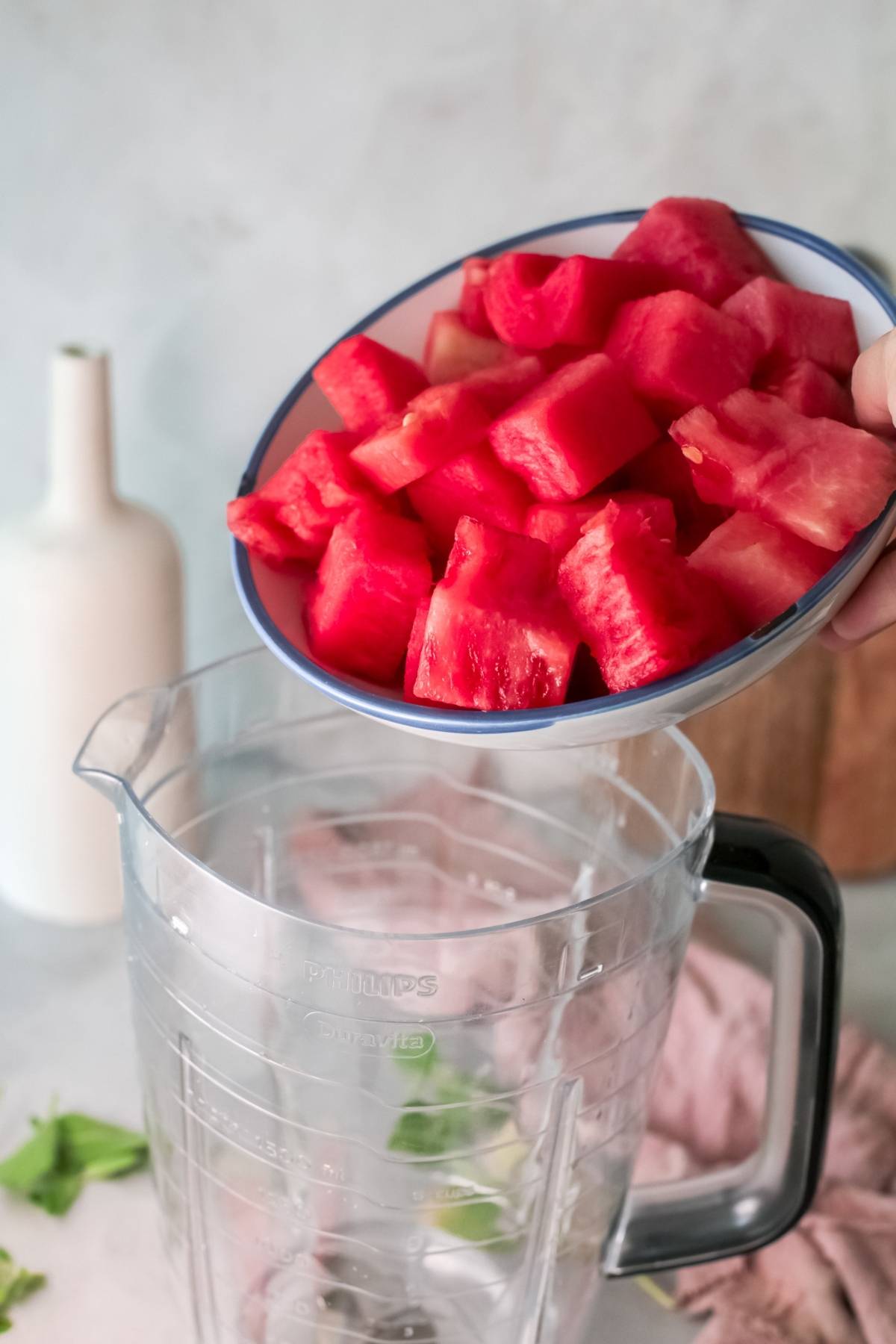 A hand pours a bowl of watermelon cubes into a clear blender pitcher on a kitchen counter, preparing to make a refreshing frozen watermelon cocktail.