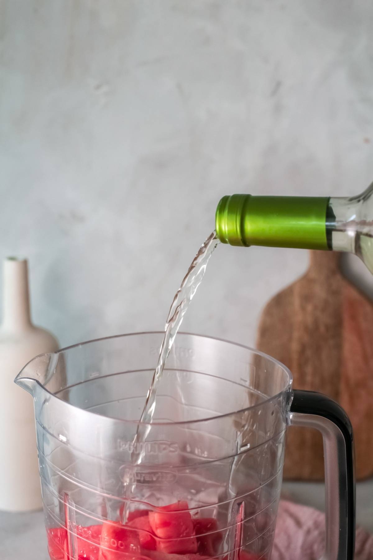 A bottle pours liquid into a pitcher filled with watermelon cubes on a kitchen counter, creating a refreshing frozen watermelon cocktail.