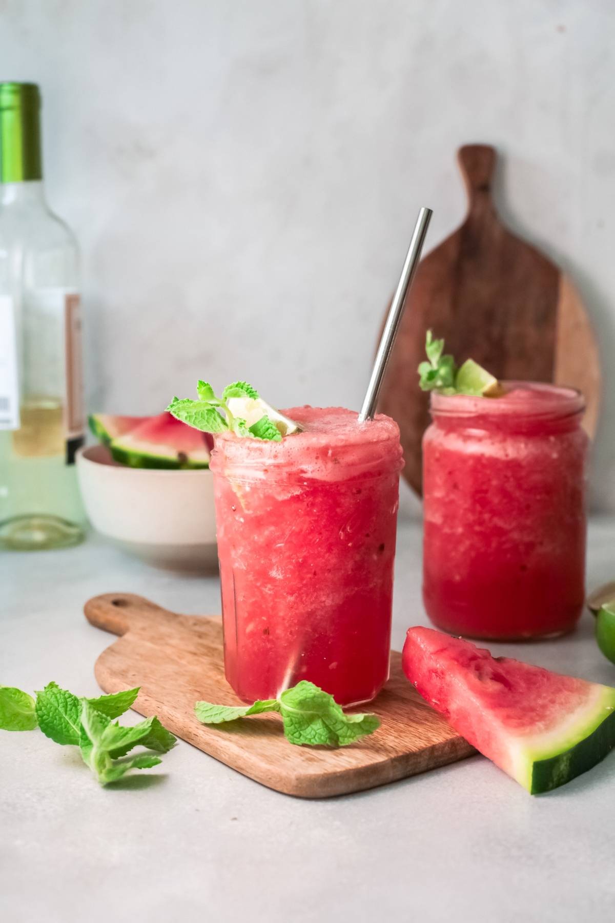 Two jars of frozen watermelon cocktail with mint and lime, served on a wooden board with fresh watermelon slices—a refreshing recipe using watermelon and wine for summer sipping.