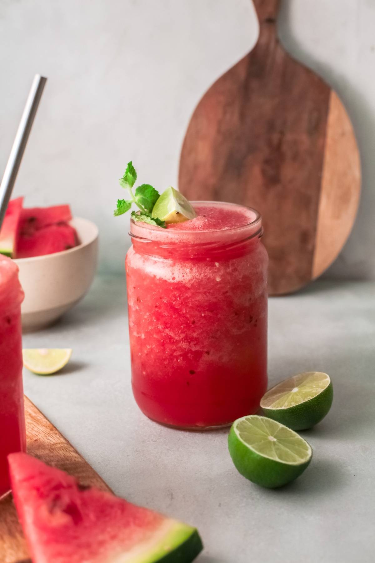 A jar of frozen watermelon cocktail smoothie with lime and mint garnish, surrounded by lime wedges and watermelon slices—perfect for a refreshing twist on a classic Pinot grigio slushie.