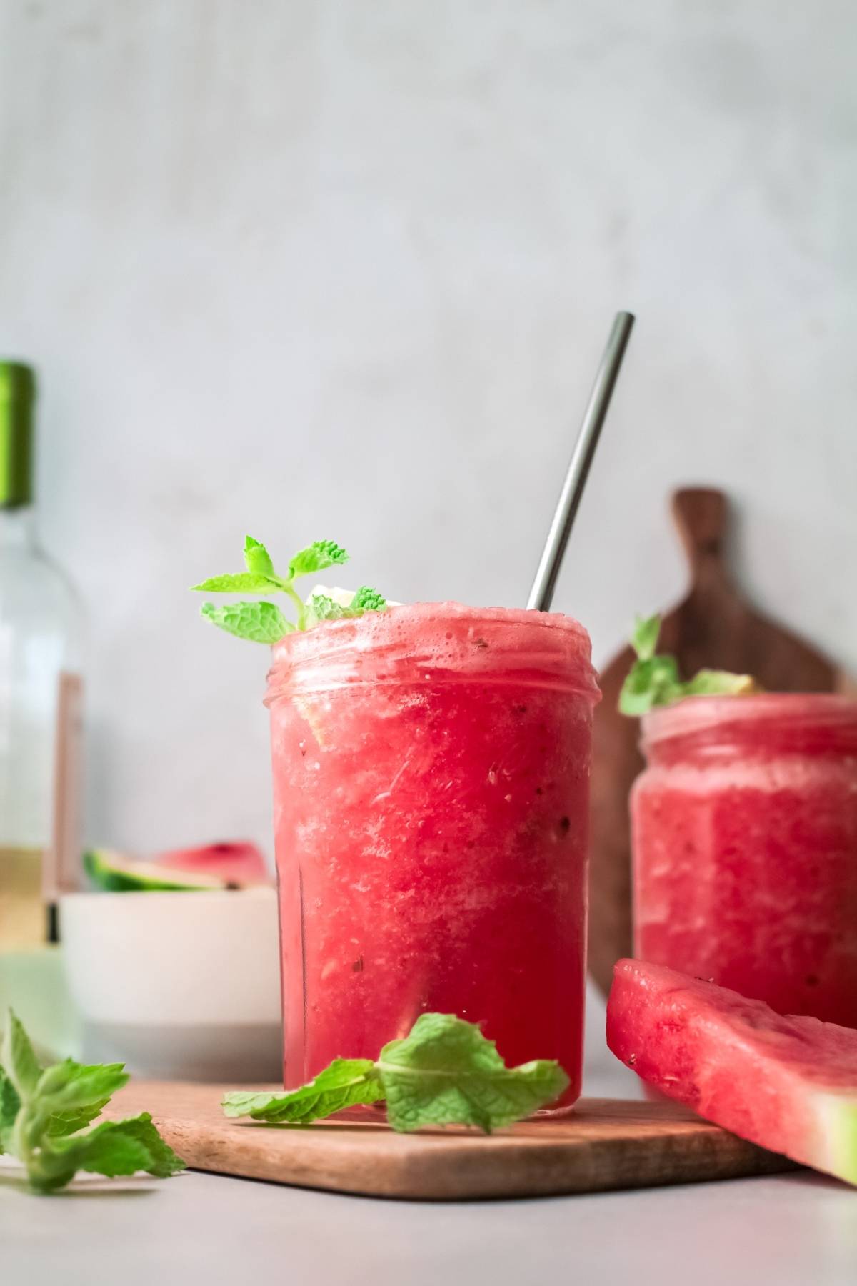 A mason jar of frozen watermelon cocktail slushie with mint leaves and a metal straw on a table, with watermelon slices nearby.