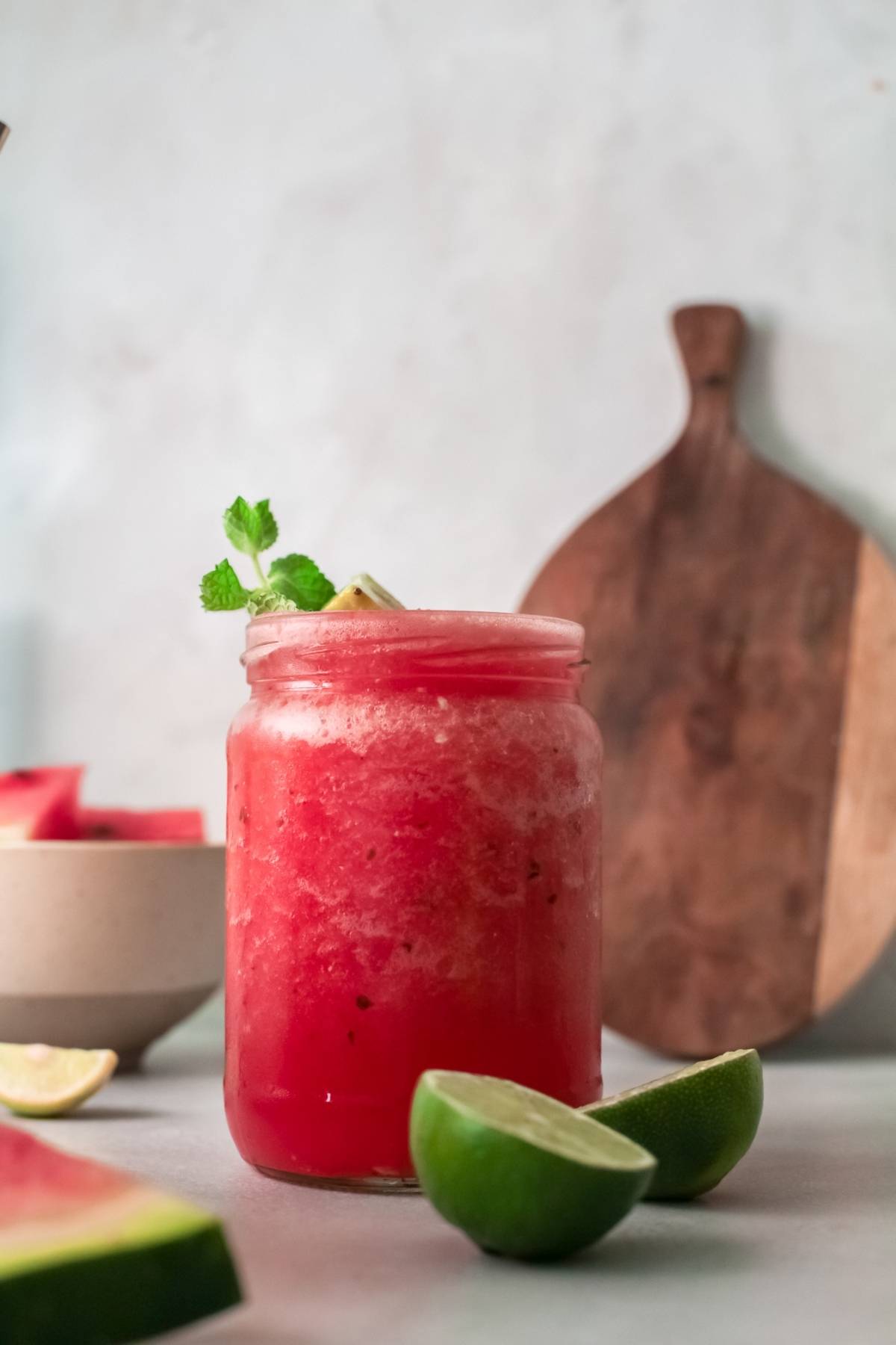 A mason jar filled with a vibrant red watermelon drink, garnished with mint, sits beside limes and watermelon slices—perfect inspiration for an alcoholic slushie drink recipe using watermelon and wine.