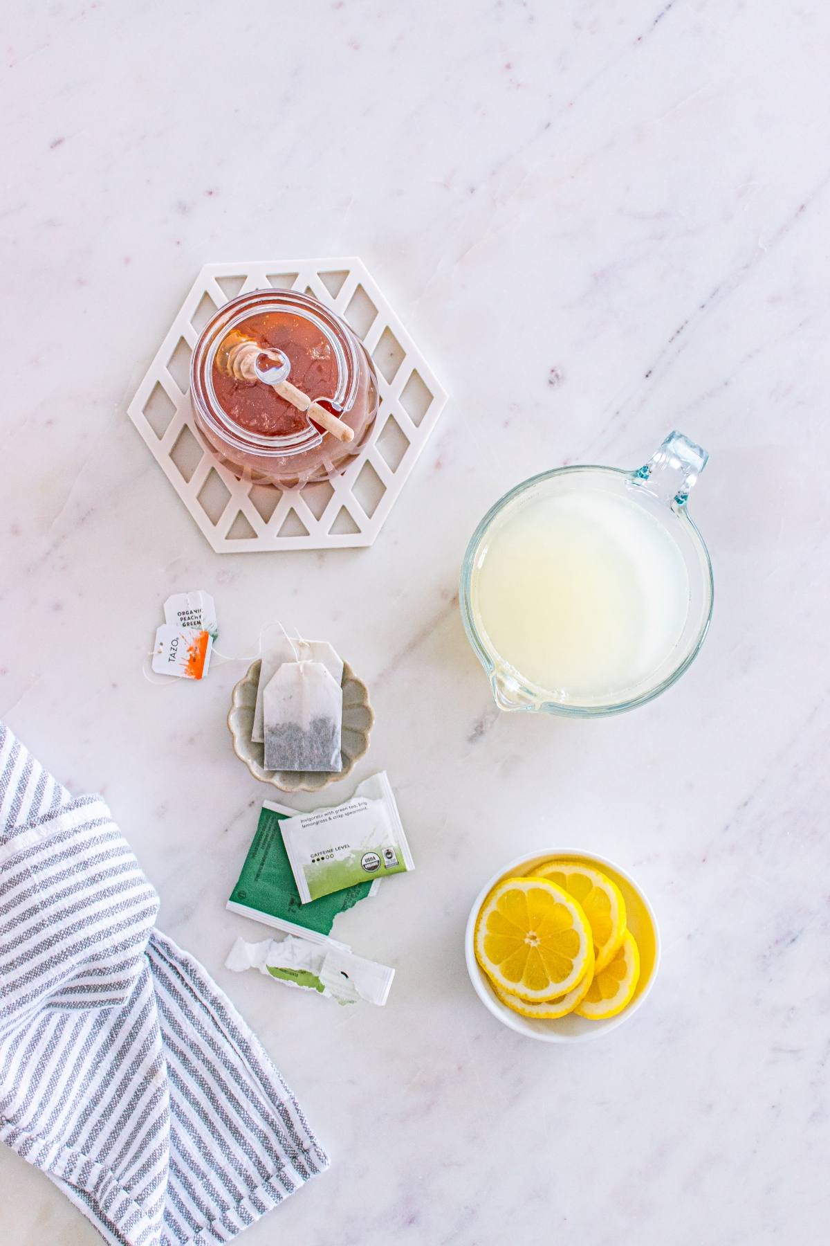Flat lay of honey jar, tea bags, lemon slices, and a glass measuring cup with lemon juice on a marble surface.