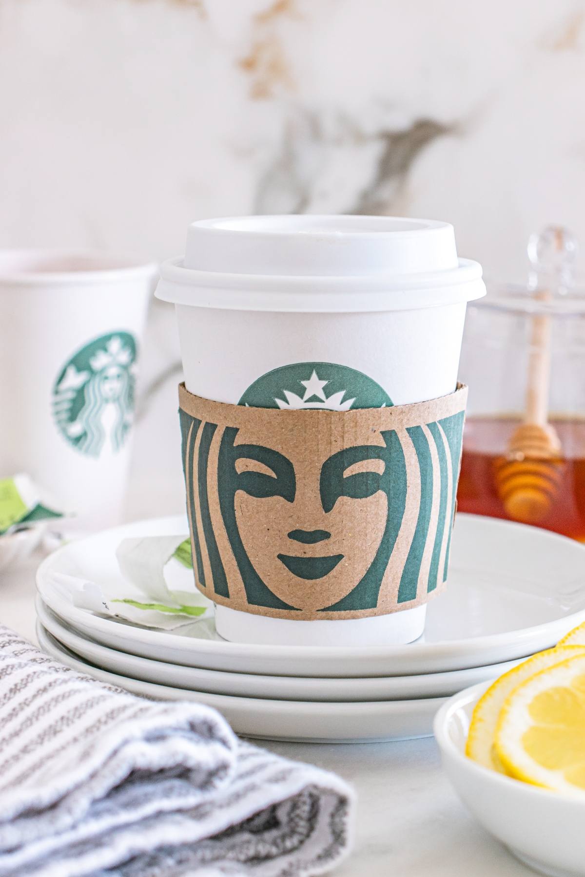 A Starbucks cup with a lid and sleeve sits on stacked plates beside lemon slices and a honey dipper.