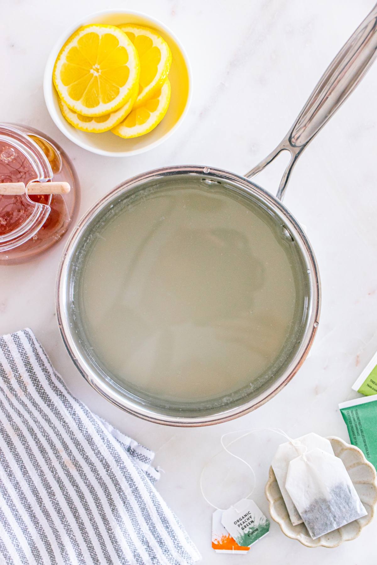 A pot of hot water, lemon slices, honey jar, tea bags, and a striped towel on a white surface.