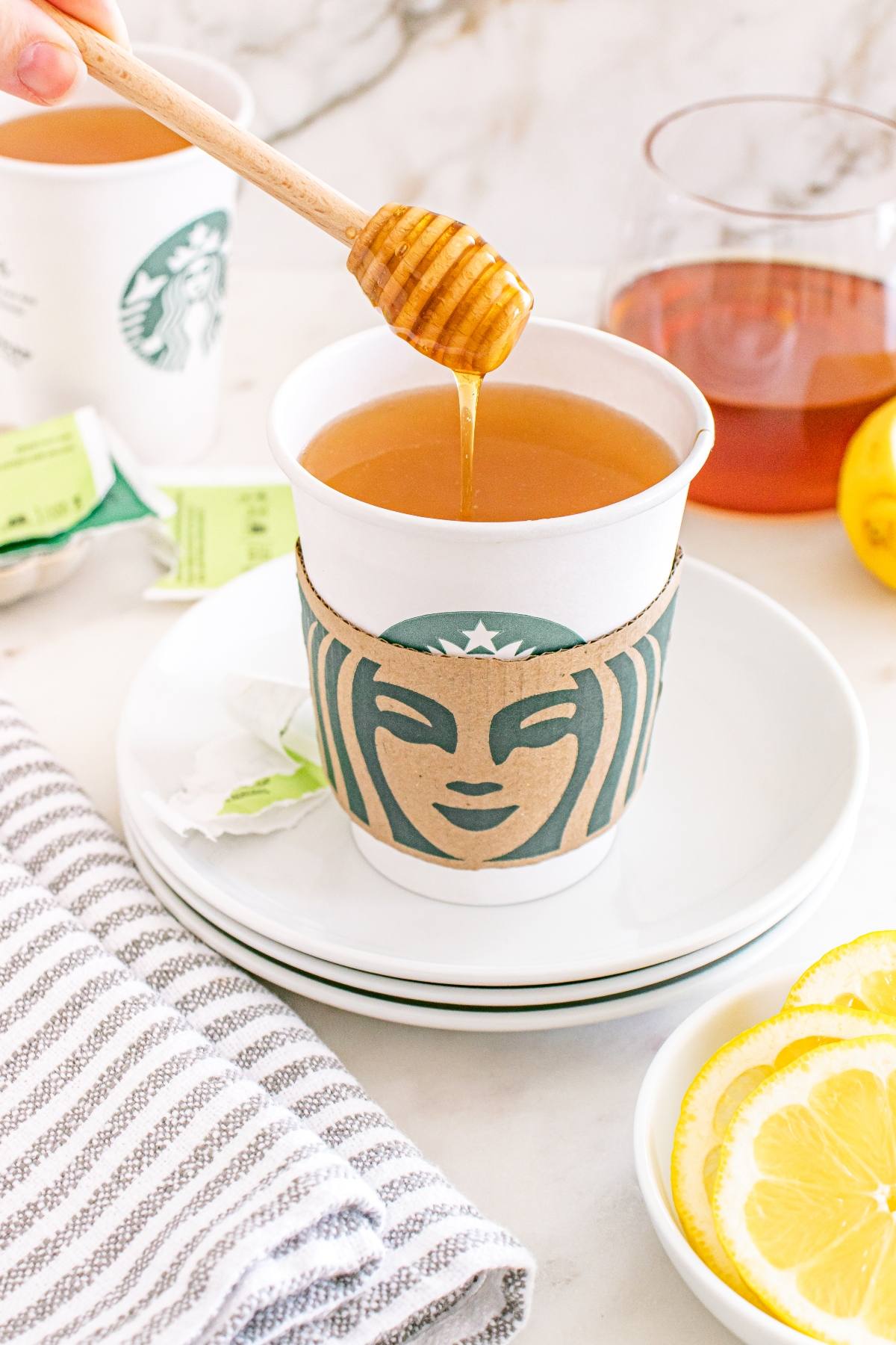 A hand drizzles honey into a Starbucks tea cup, with lemon slices and tea bags nearby.