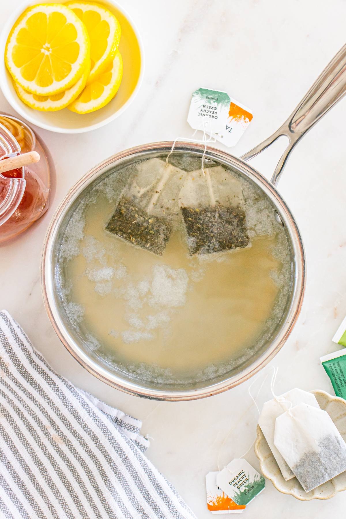A saucepan with tea bags steeping in hot water, surrounded by lemon slices, honey, and a striped towel.