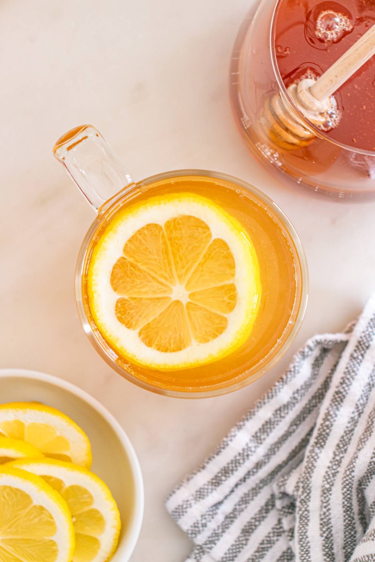A glass cup of tea with a lemon slice, honey dipper in jar, lemon slices, and striped towel on table.