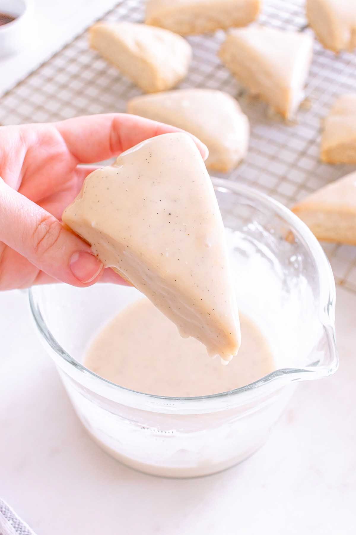 Hand dipping a triangle Starbucks Copycat Vanilla Bean Scone into white glaze, with more scones cooling on a rack in the background.