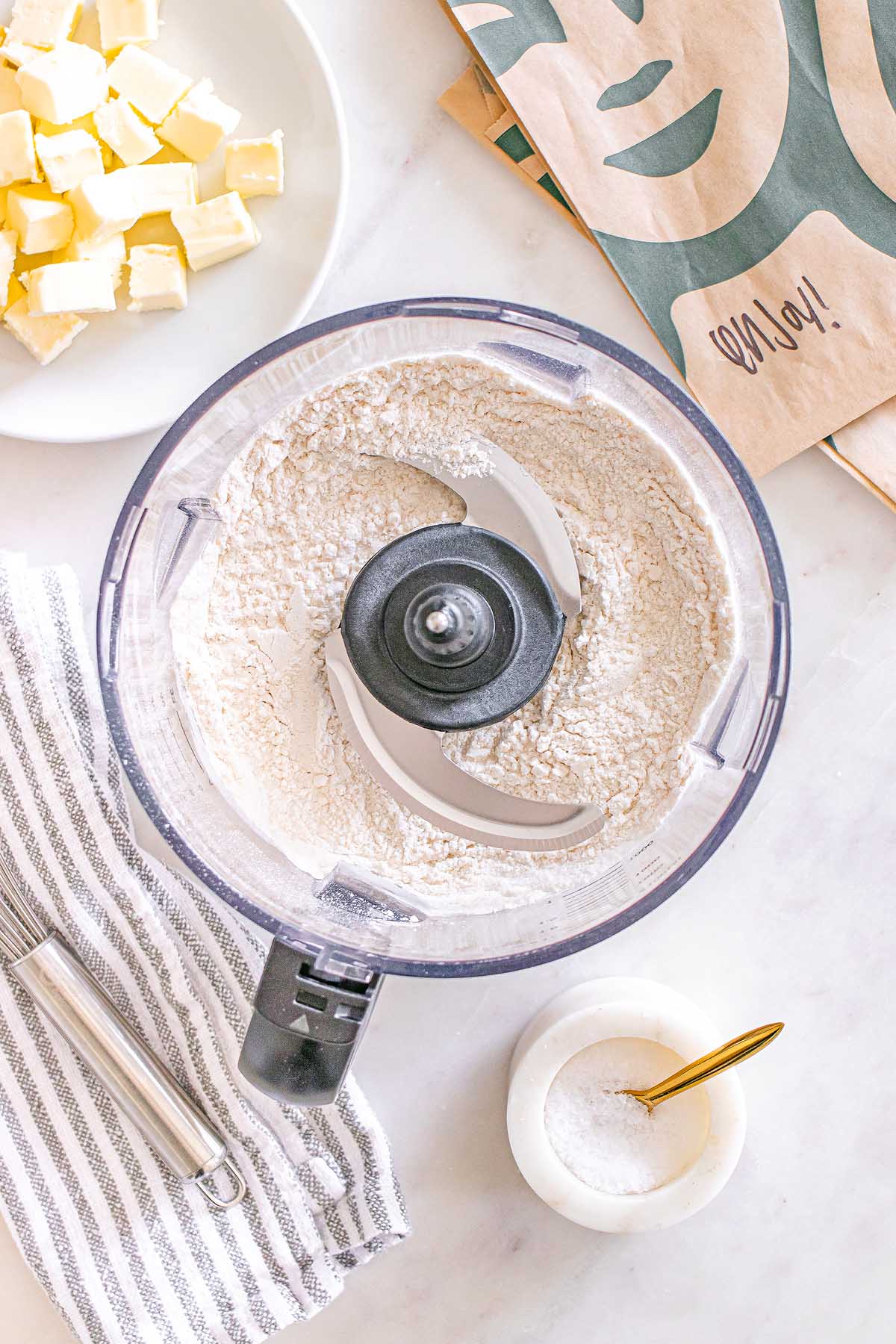 Food processor with flour inside, butter cubes on a plate, and a small bowl of salt on a countertop—everything ready to make Starbucks Copycat Vanilla Bean Scones.